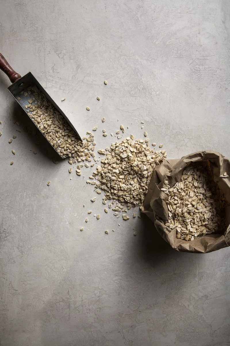 Oat flakes spilled from a trowel onto a gray surface, with some flakes inside a paper bag.
