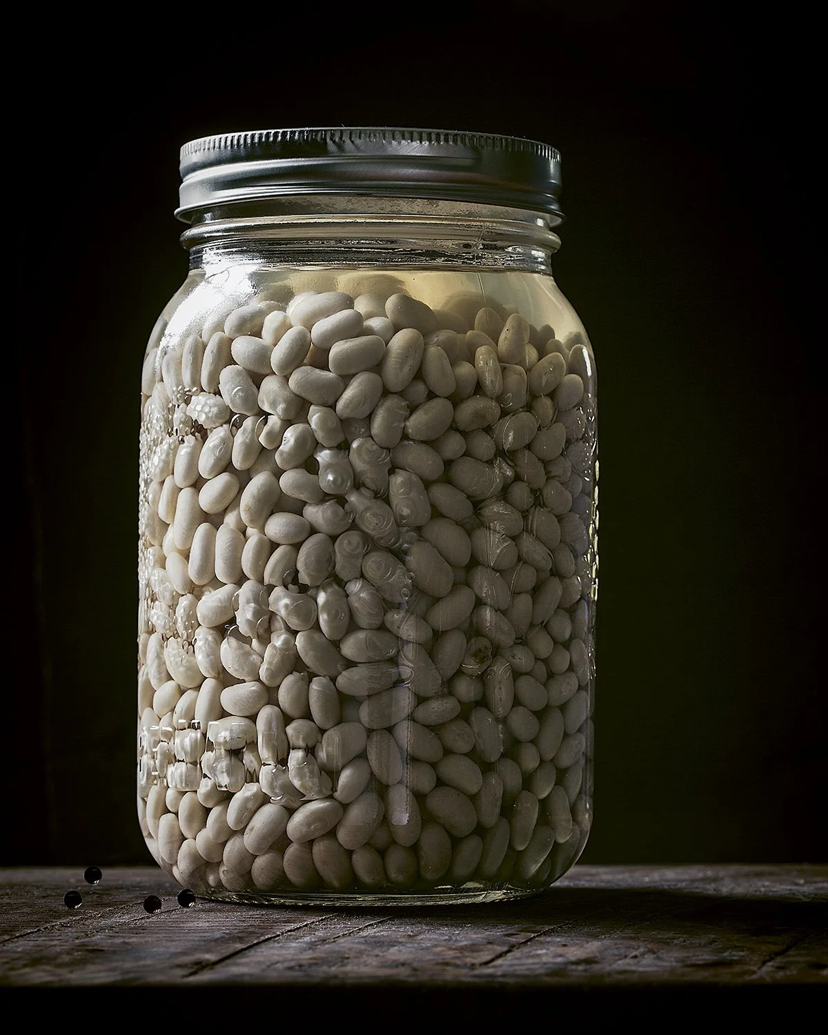 A glass jar filled with white beans sitting on a wooden surface against a dark background.