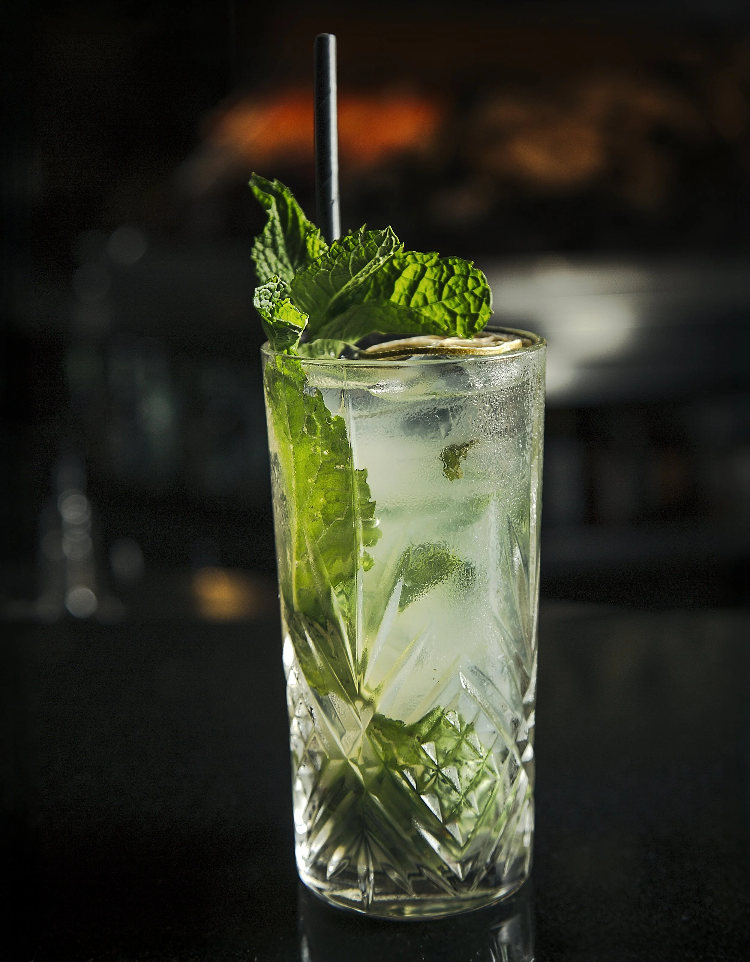 A tall glass of mojito with mint leaves, lime wedges, ice, and a black straw on a dark surface.