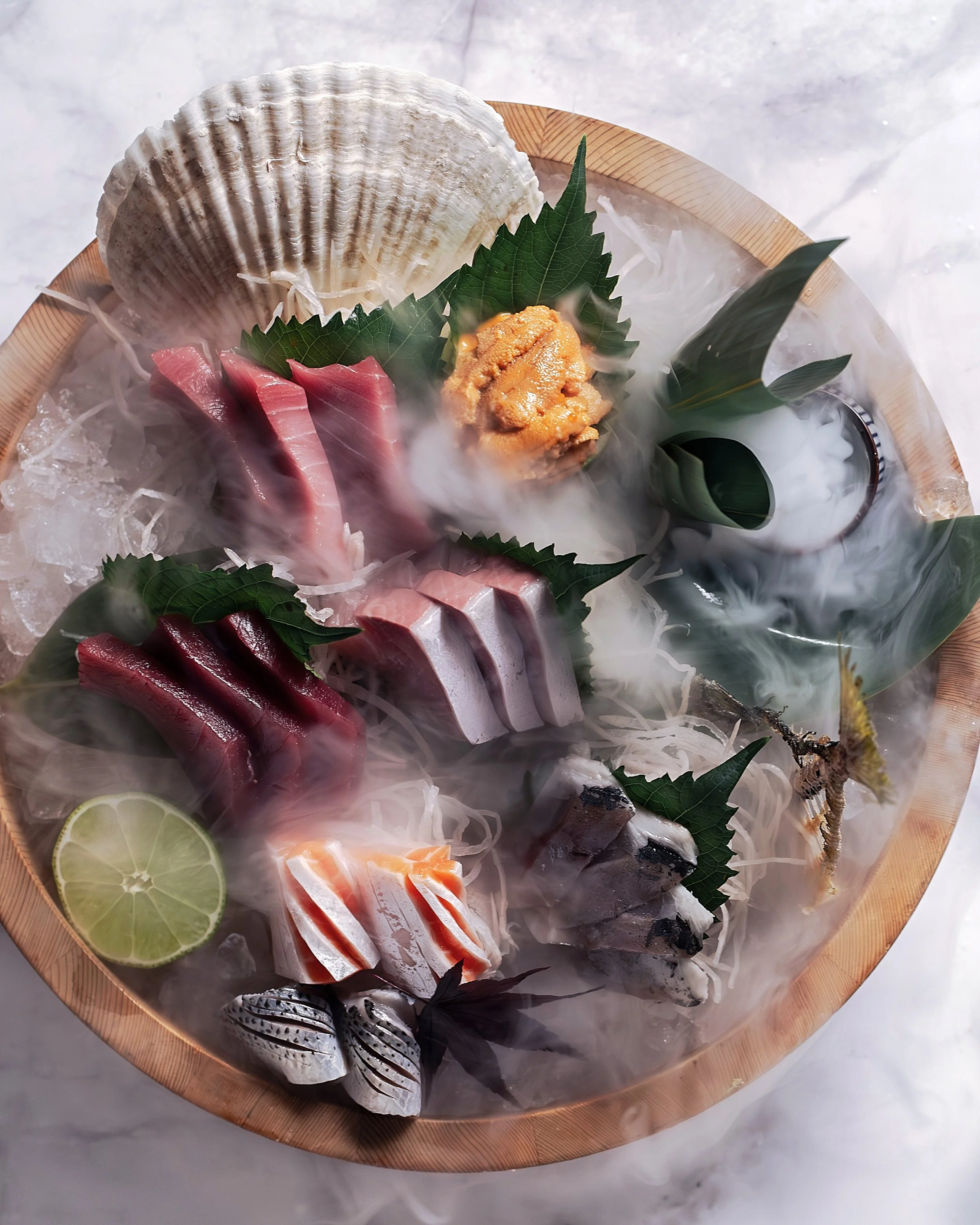 Sashimi platter with various slices of raw fish, garnished with green leaves, a lime wedge, and dry ice fog creating a smoky presentation.
