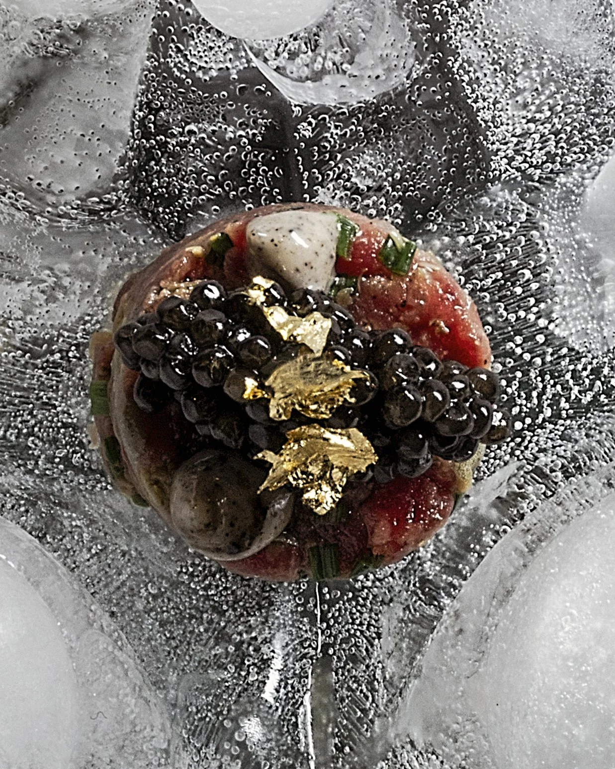A dish with black pearls, green herbs, a dollop of white sauce or mayonnaise, garnished with gold leaf, on a bed of red and green vegetables or ingredients, all presented in a transparent container with bubbles and water.