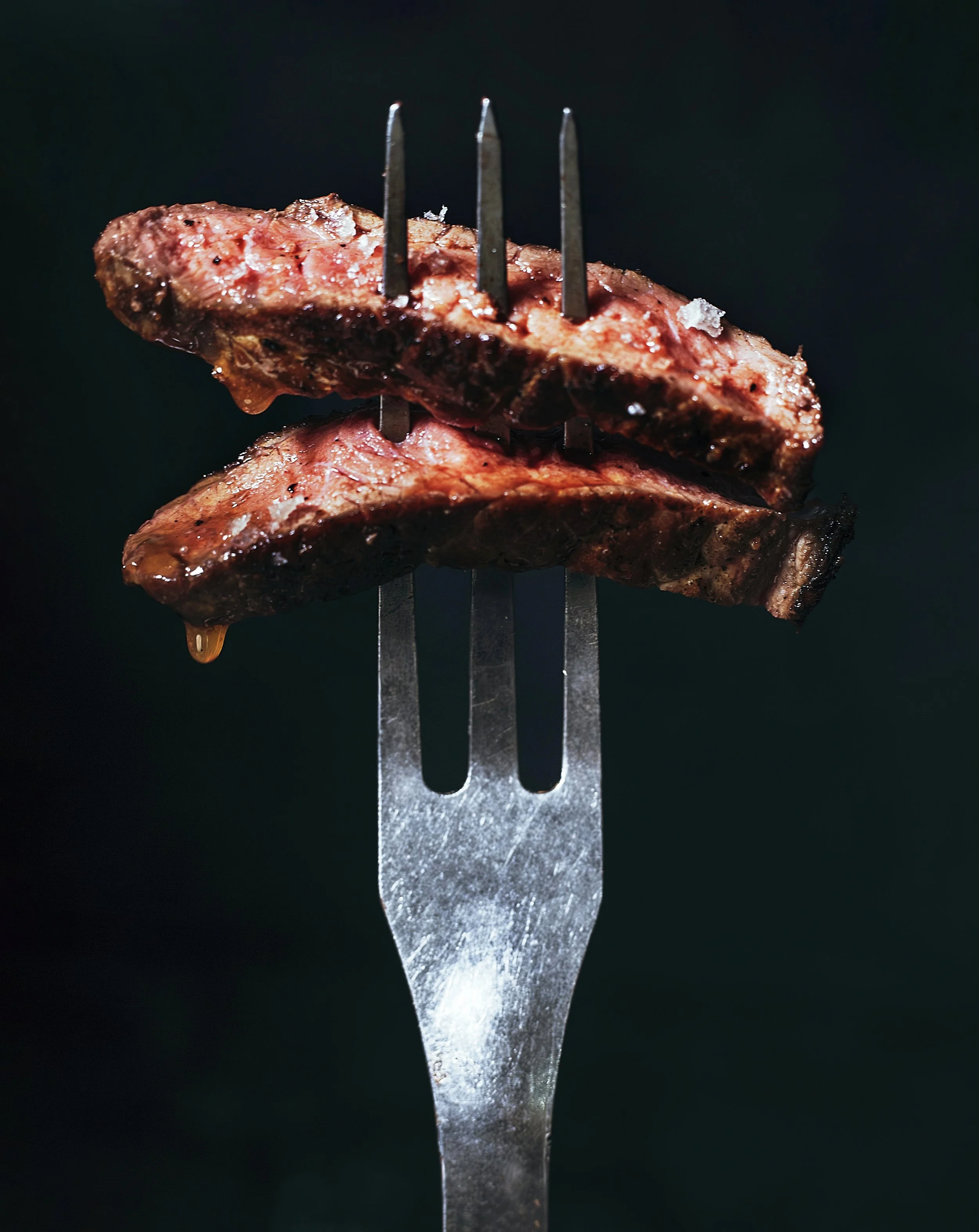 Close-up of a fork with two grilled pieces of steak against a dark background.