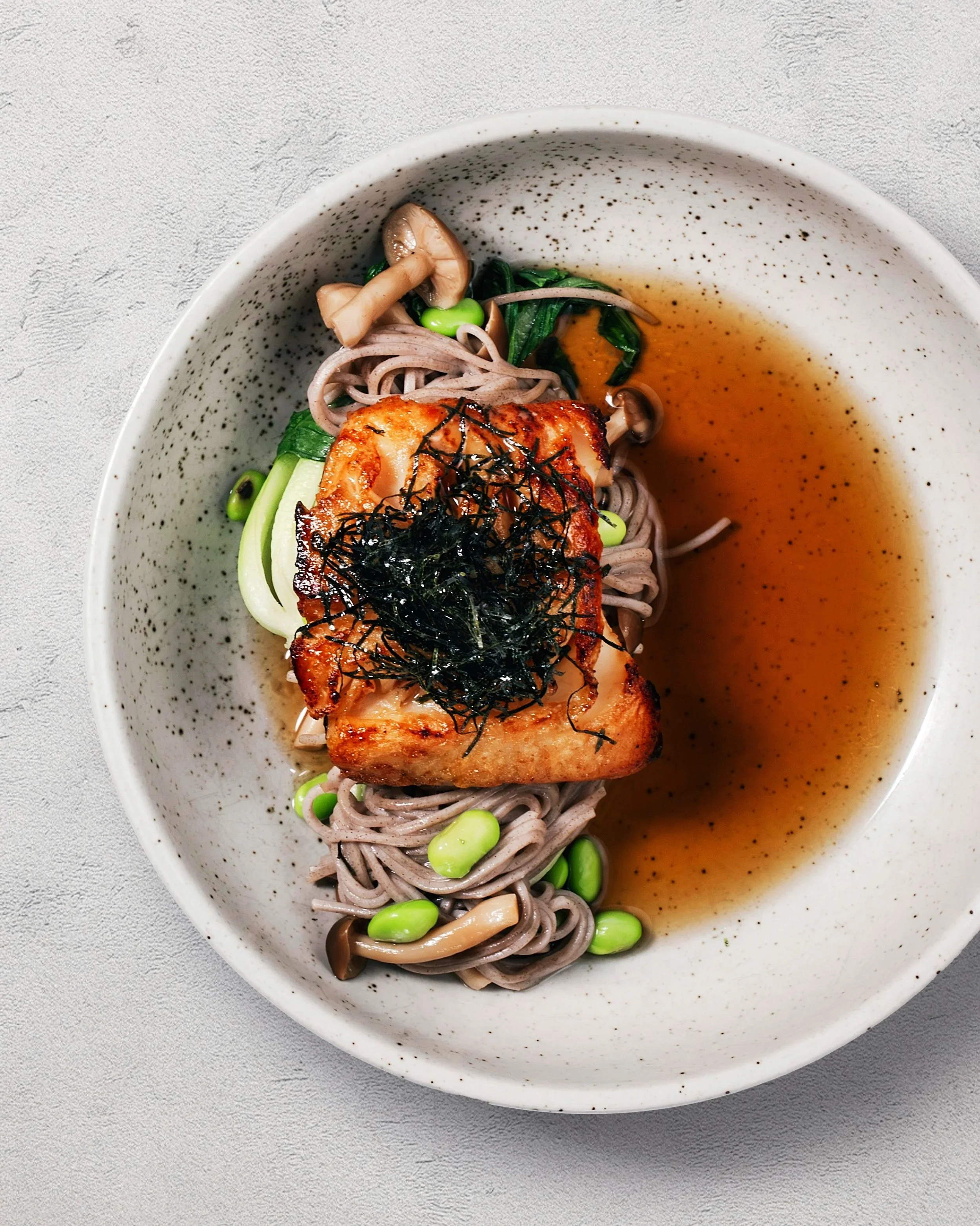 Bowls of soba noodles with grilled fish, edamame, mushrooms, greens, and broth garnished with shredded nori
