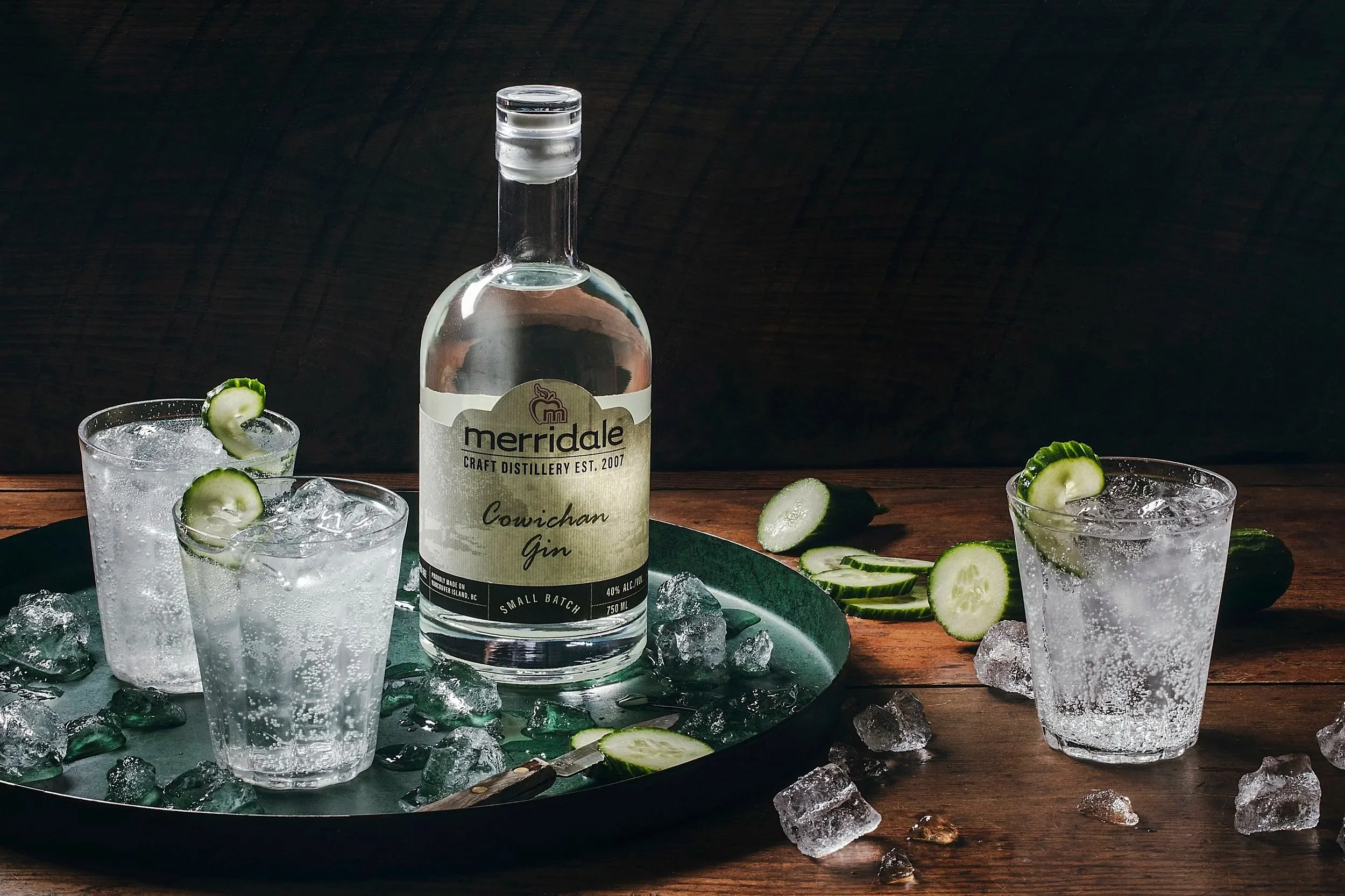 A bottle of Merridale Cowichan Gin on a tray with two glasses filled with gin and cucumber garnish, surrounded by cucumber slices, ice cubes, and a wooden surface.