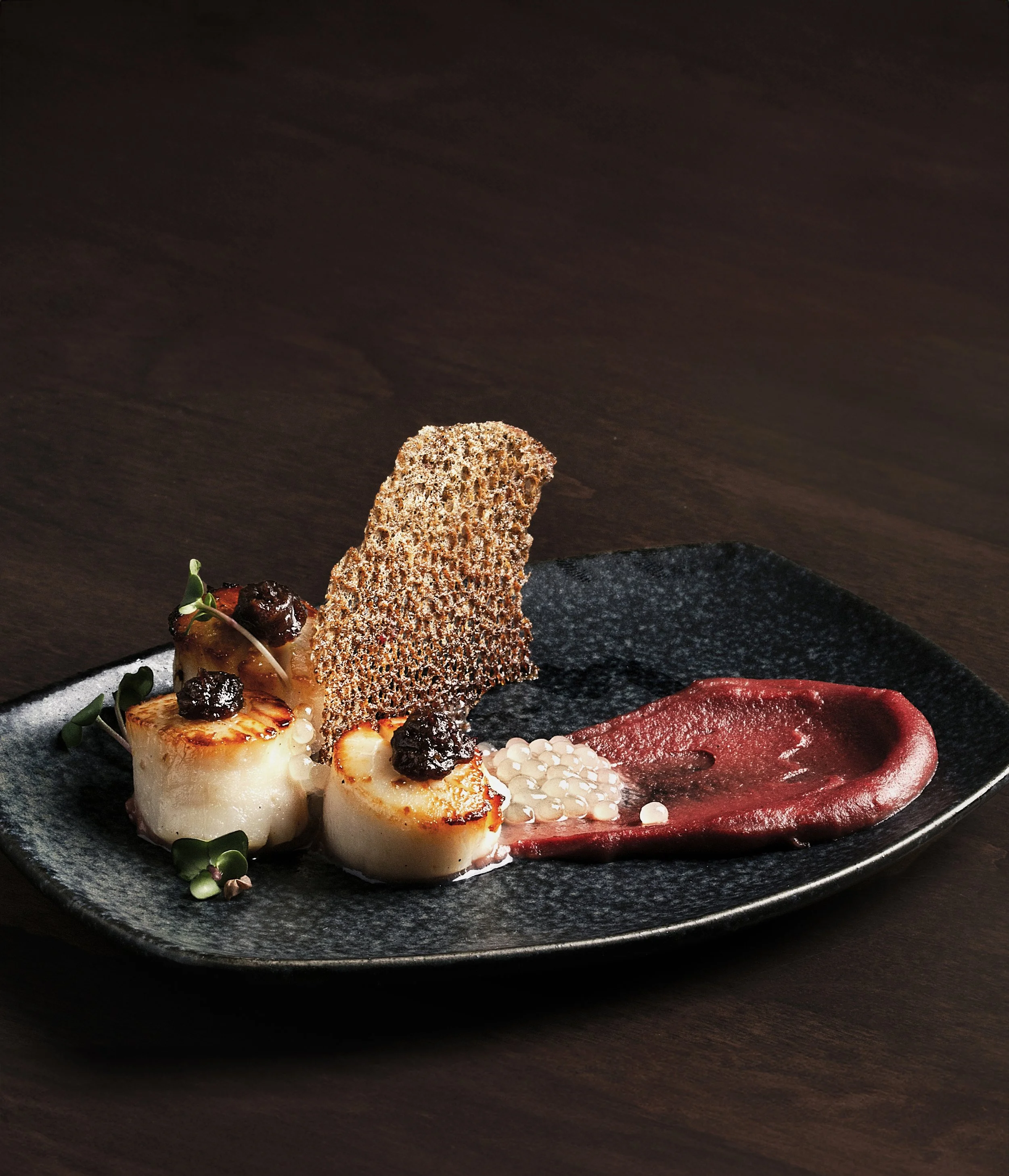 A gourmet dish featuring seared scallops with blackberries, garnished with microgreens and a delicate, crisp tuile on a black plate.