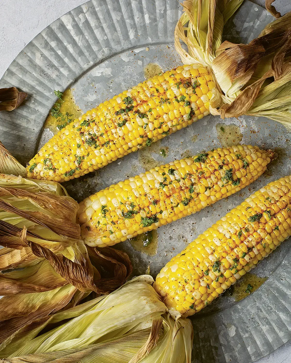 Three ears of grilled corn on a metallic tray with husks partially peeled back.