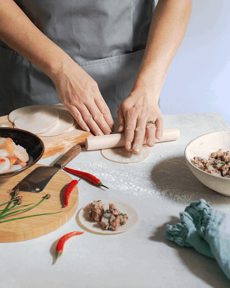Person rolling out dough with a rolling pin on a kitchen counter, with bowls of filling, a chopping knife, and chili peppers nearby.