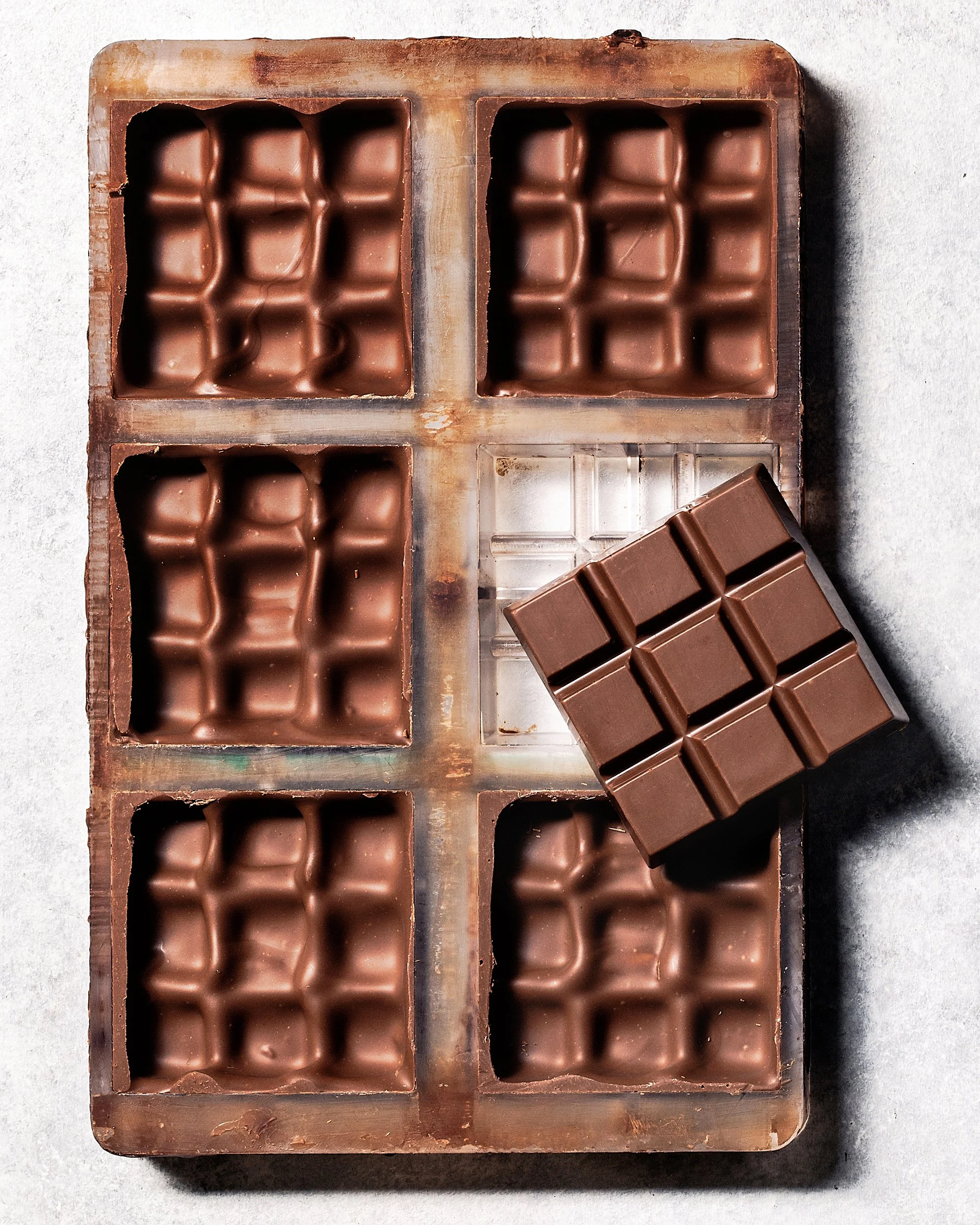 A vintage chocolate mold with eight cavities, five filled with chocolate, one empty, and a chocolate bar resting on it.