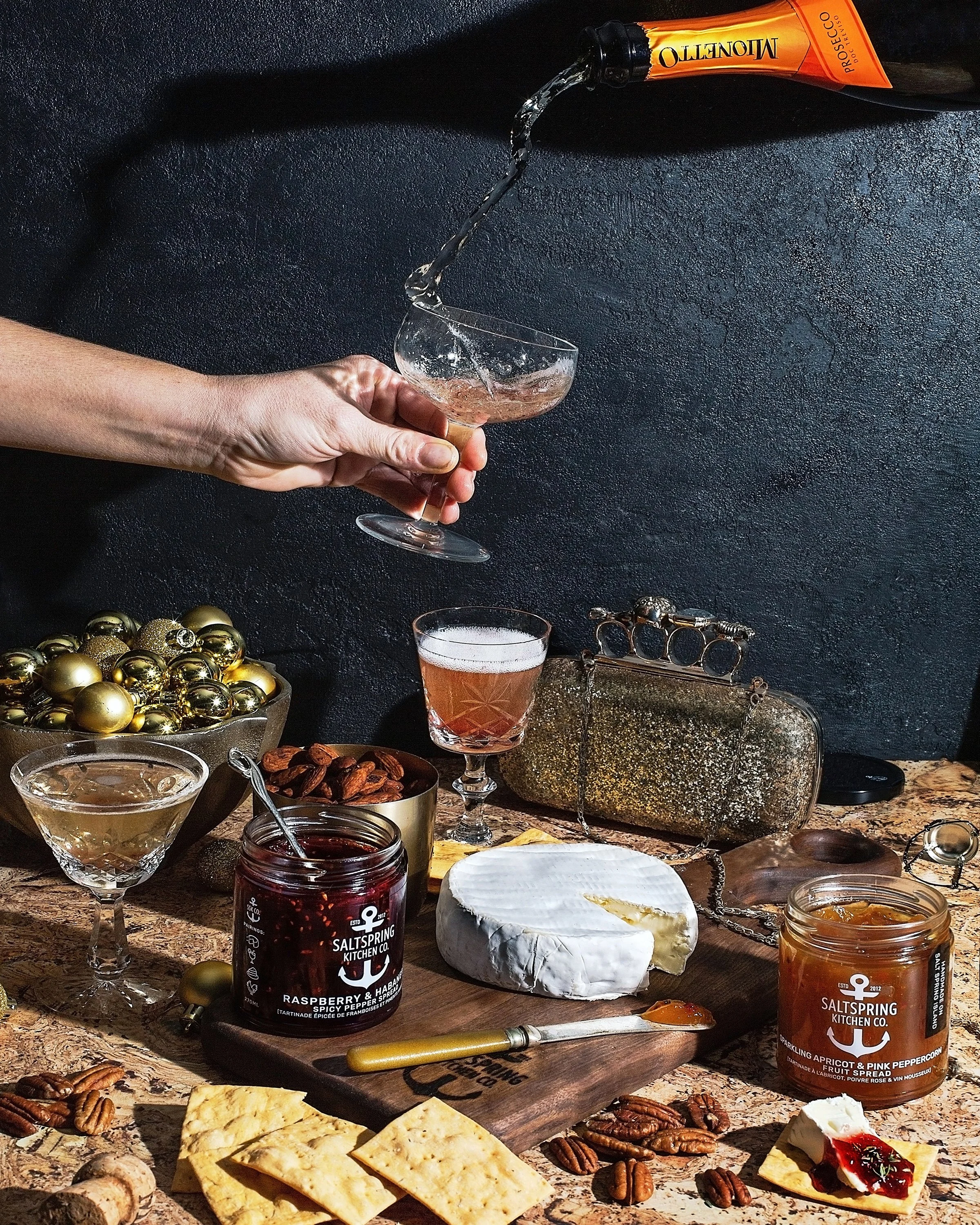 A woman pouring champagne into a coupe glass above a table with assorted cheeses, jams, crackers, nuts, and decorative items, set against a dark textured background.