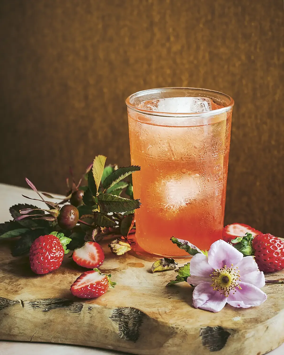 A glass of pink strawberry-infused beverage with ice on a wooden surface, surrounded by fresh strawberries, strawberry leaves, and a pink flower, with a dark brown background.