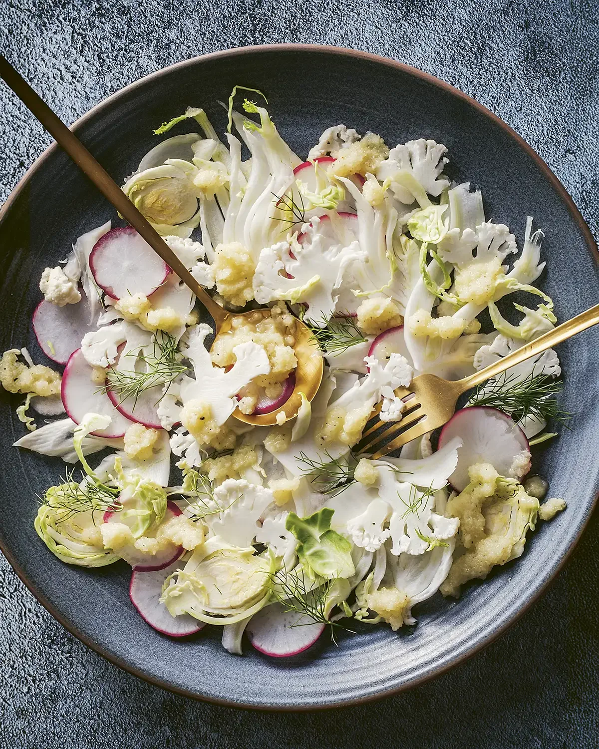 A plate of cauliflower, radish slices, and fennel garnished with dill, with a gold fork and spoon.