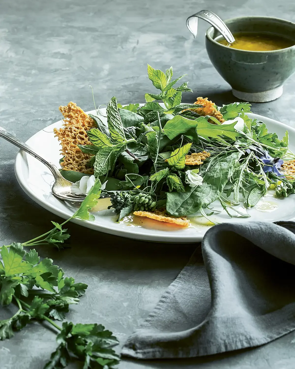 A plate of fresh mixed greens with herbs, crispy bread crisps, and edible flowers, accompanied by a bowl of dressing or soup on a gray surface with a gray napkin.