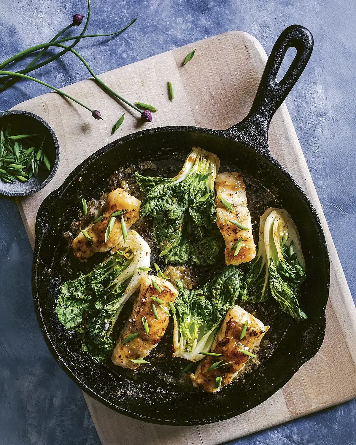 Skinless chicken breasts with sage and garlic roasting in a cast iron skillet with whole and halved fennel bulbs, garnished with chopped green herbs, on a cutting board.