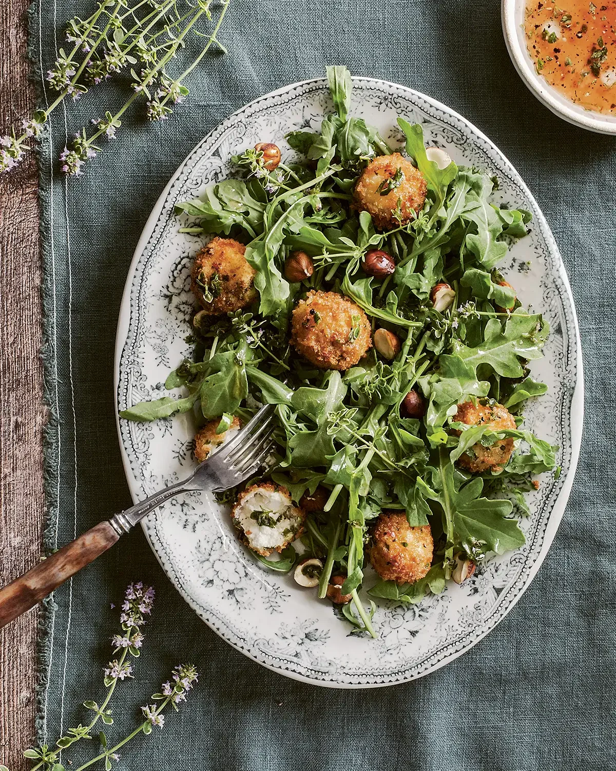 A plate of arugula salad with breaded, fried cheese balls and mixed nuts, on a floral-patterned white platter, with a fork resting on the plate. There is a bowl of orange soup and some lavender sprigs on the table.