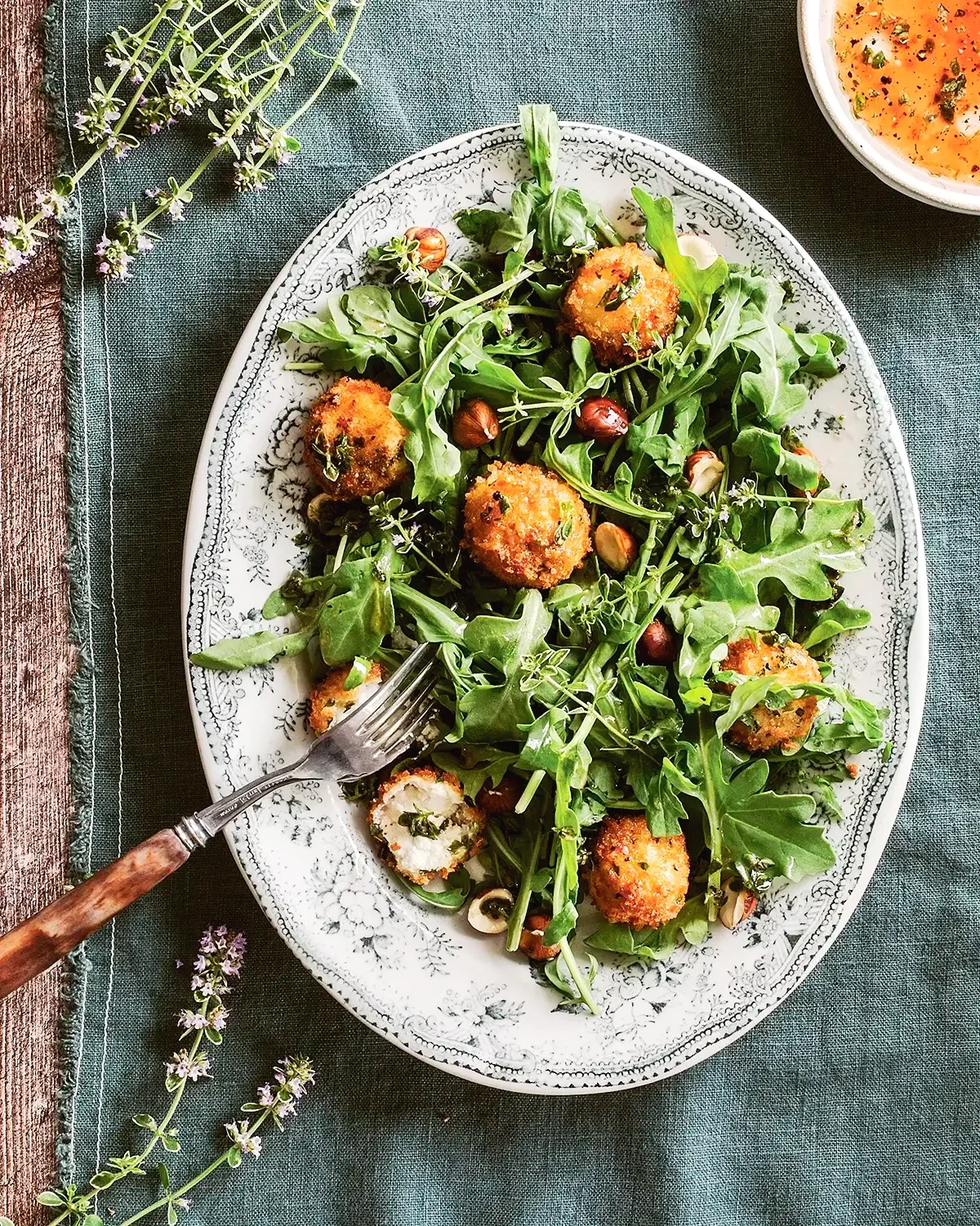 A white oval plate filled with arugula and mixed greens salad topped with breaded and fried cheese balls and garnished with herbs.