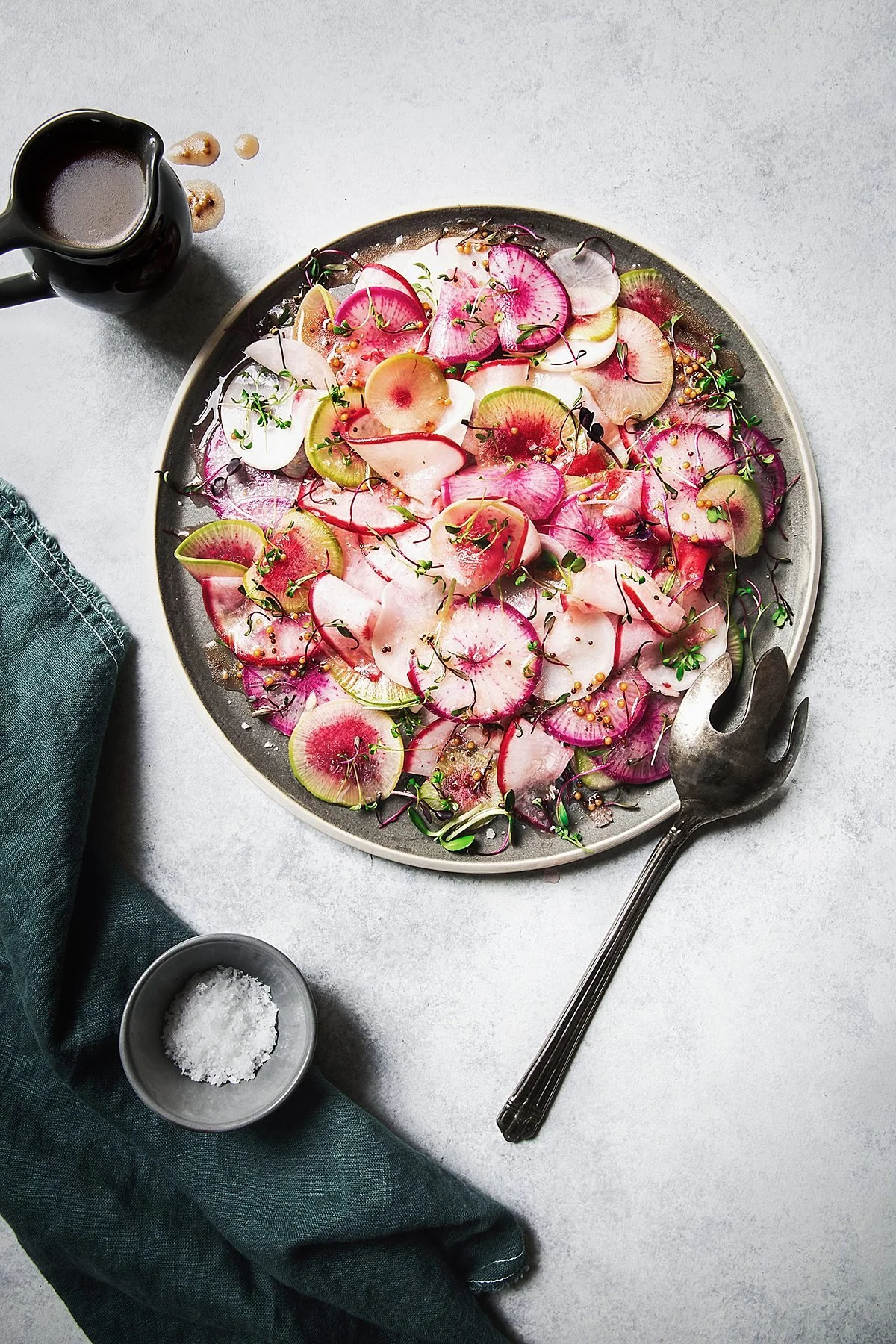 A round plate with thinly sliced radishes, garnished with herbs and mustard seeds, on a light gray surface with a black salad fork, a small bowl of coarse salt, a small bowl of salad dressing, a folded dark green cloth, and a small pitcher of dressin