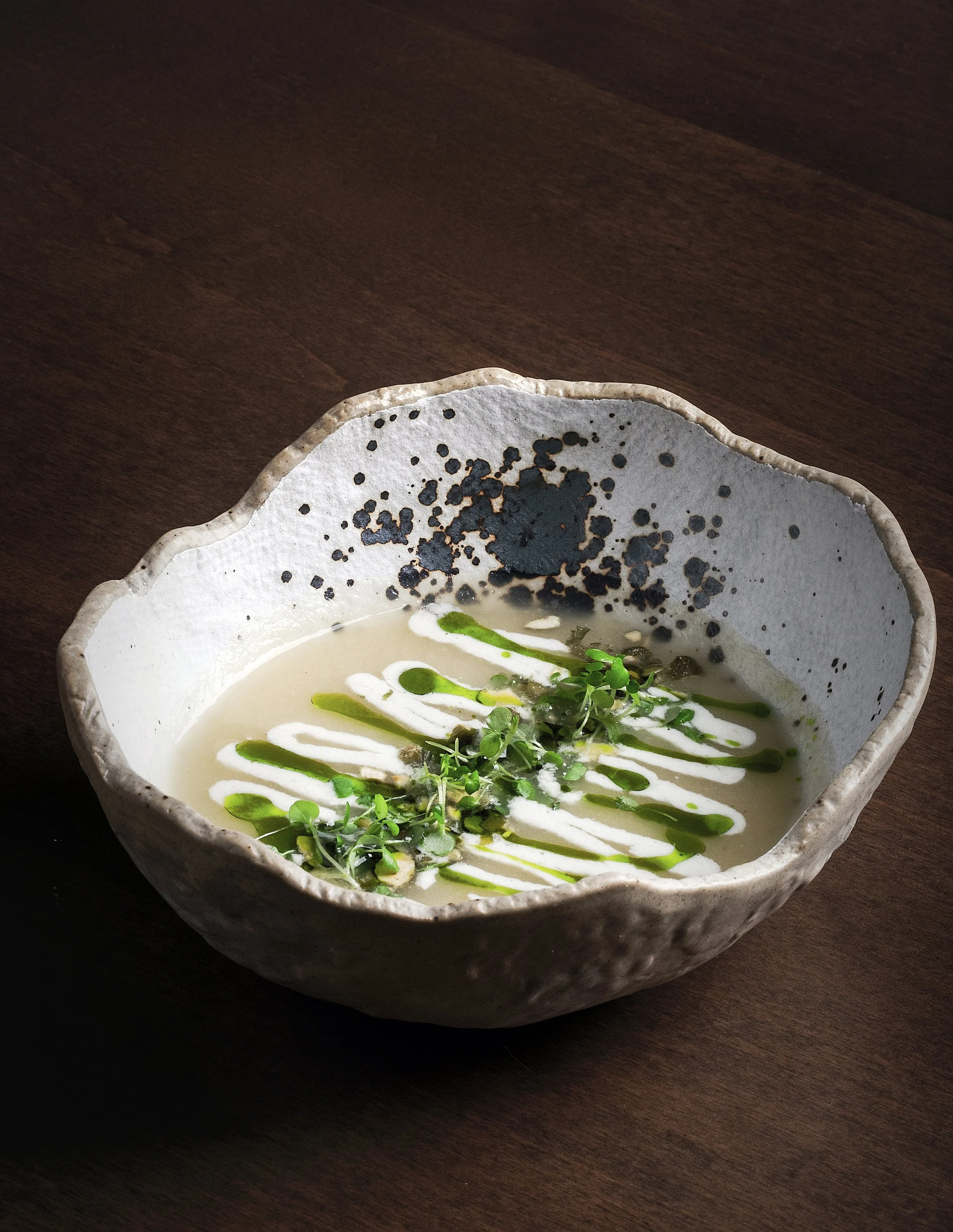 A ceramic bowl containing a creamy soup garnished with microgreens, drizzled with white and green sauces, placed on a dark wooden surface.