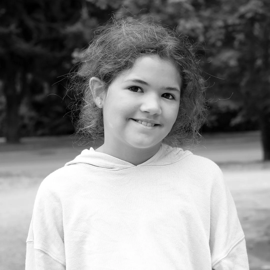 Black and white photo of a young girl with curly hair smiling outdoors, wearing a light-colored hoodie.