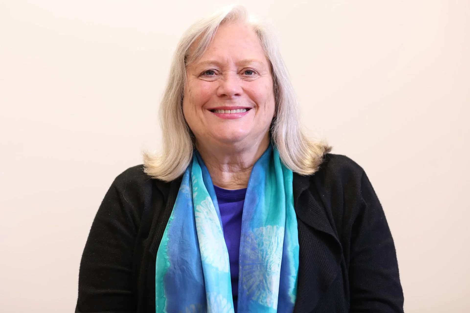 Smiling woman with shoulder-length gray hair wearing a black sweater and a blue patterned scarf, against a plain light-colored background.