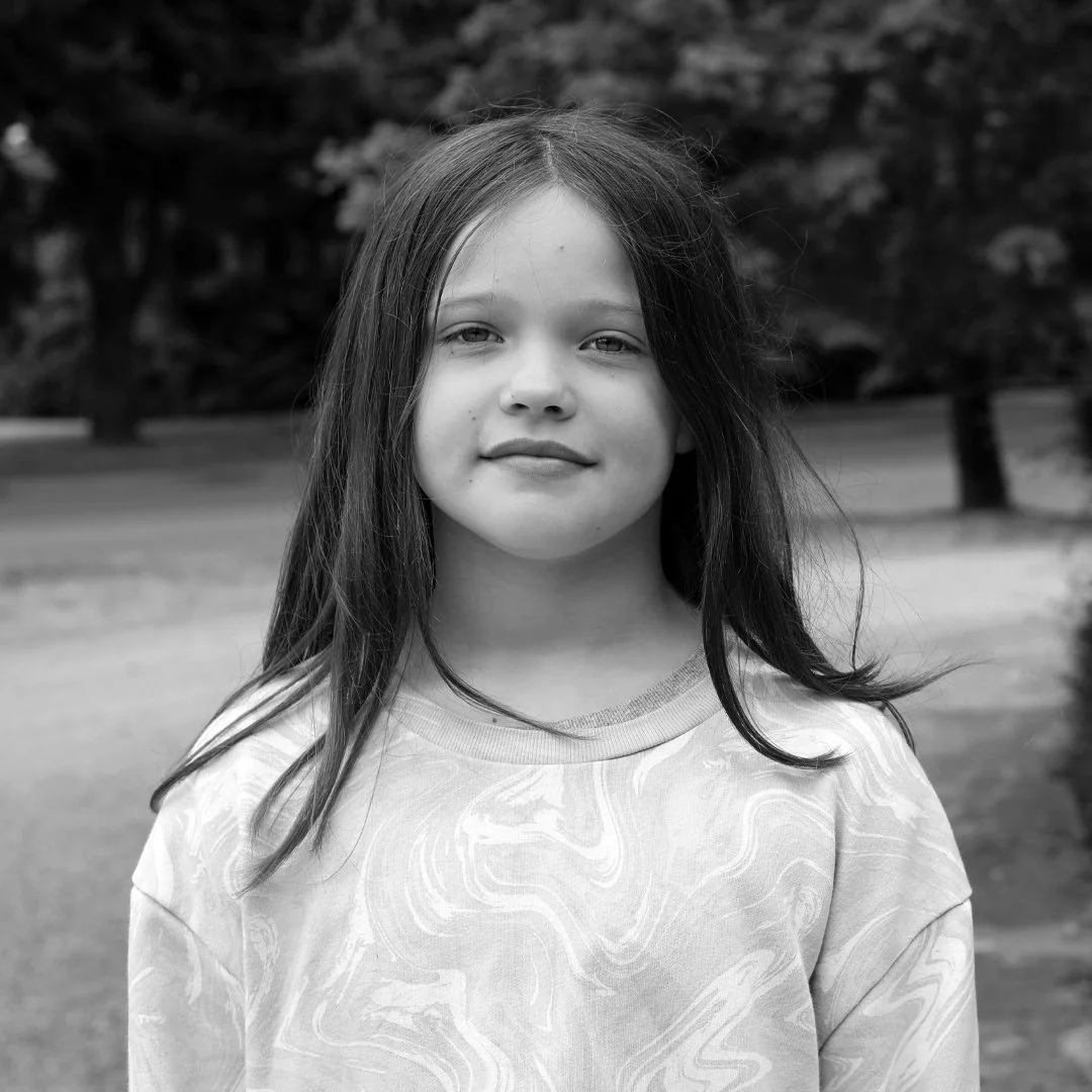 A young girl with long hair standing outdoors in front of trees, wearing a patterned shirt, looking at the camera with a slight smile.