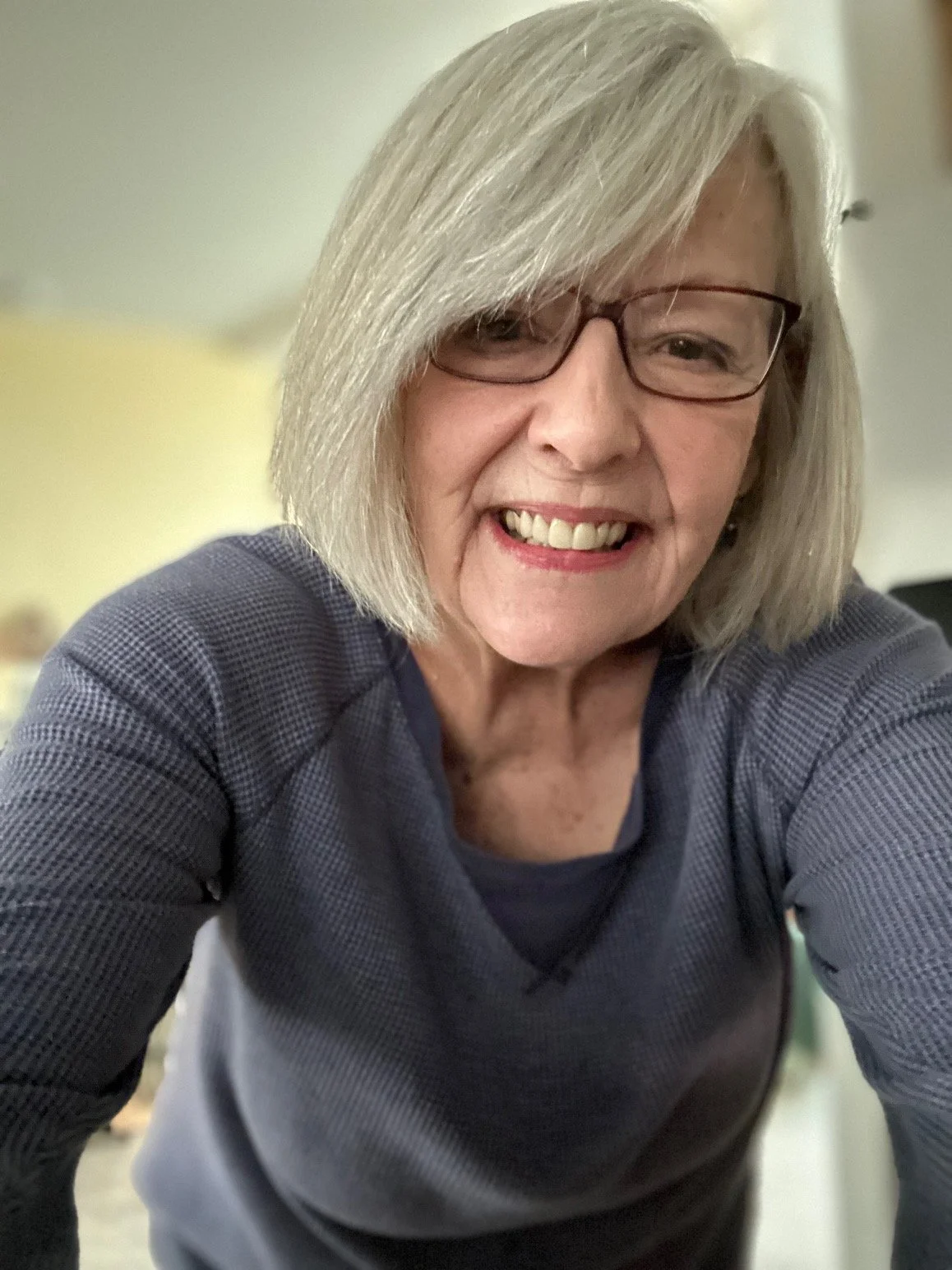 Close-up of a smiling elderly woman with gray hair, glasses, wearing a dark blue shirt, taking a selfie indoors.