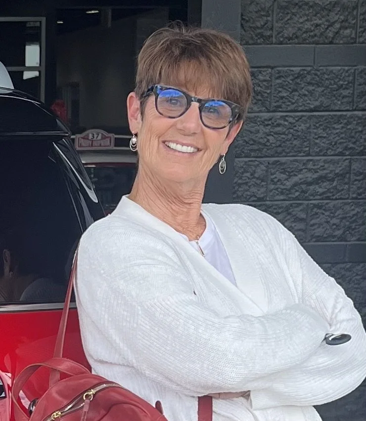 A smiling woman with short brown hair wearing glasses, a white sweater, and earrings, standing in front of a dark textured wall and a black car.