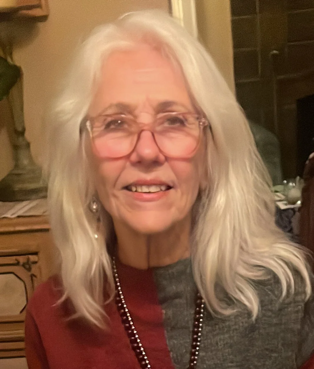 A smiling woman with long white hair, wearing glasses, a red and gray top, and a beaded necklace, sitting indoors.