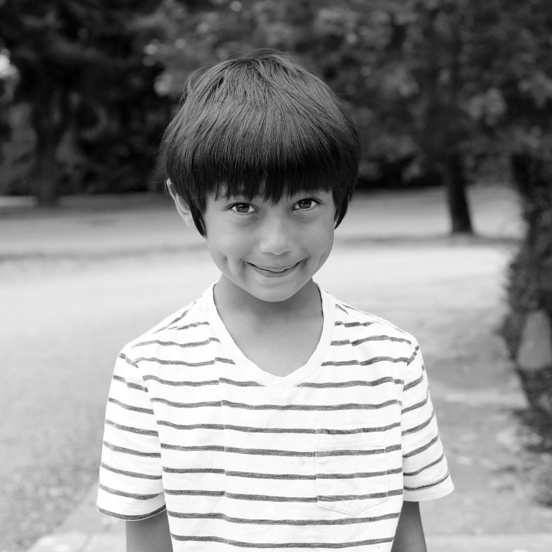 A young boy with a short, dark hair styled in a bowl cut, smiling slightly, standing outdoors in a park, wearing a striped t-shirt, in black and white.