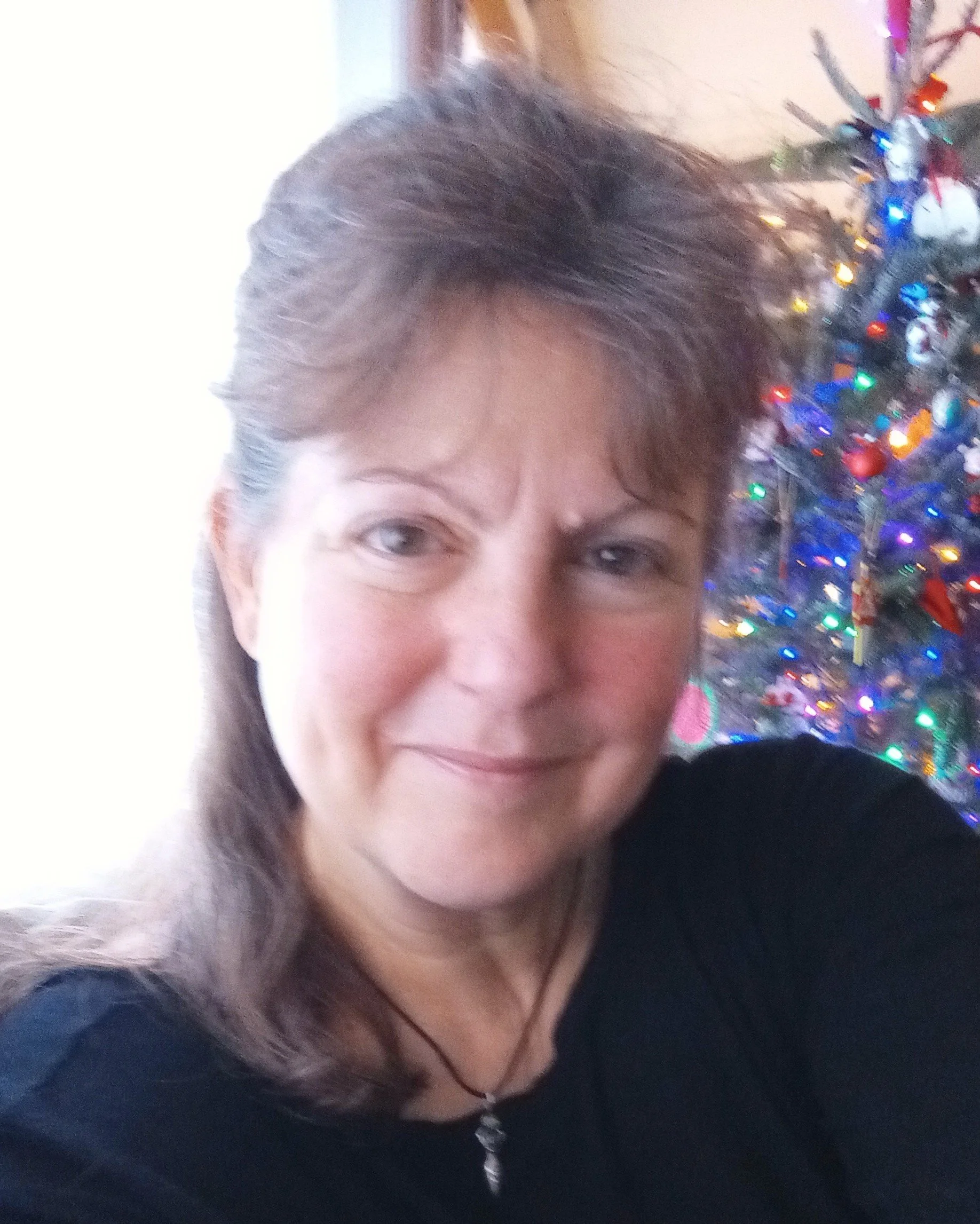 A woman with brown hair and a black top taking a selfie in front of a brightly lit Christmas tree.