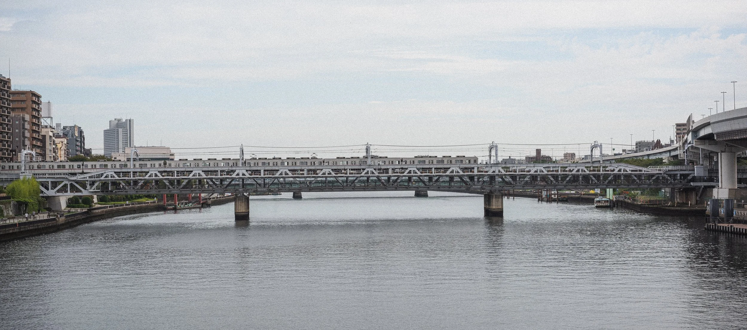 A cityscape featuring a river with a bridge and train tracks crossing over it. Buildings are visible in the background, with some greenery along the riverbank.