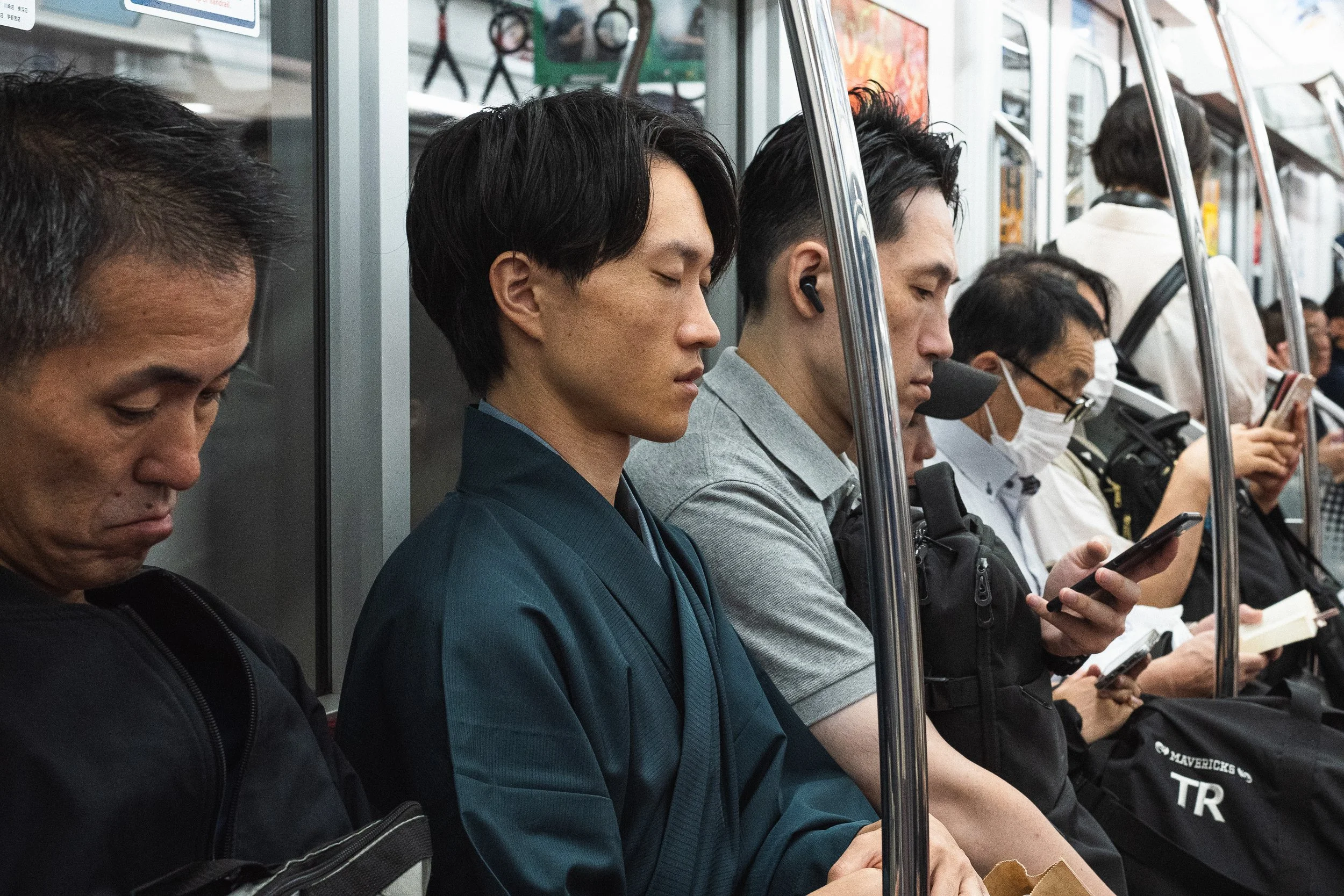 People sitting on a subway train, most looking at their phones, including a man wearing a mask and glasses, with a diverse group of passengers.