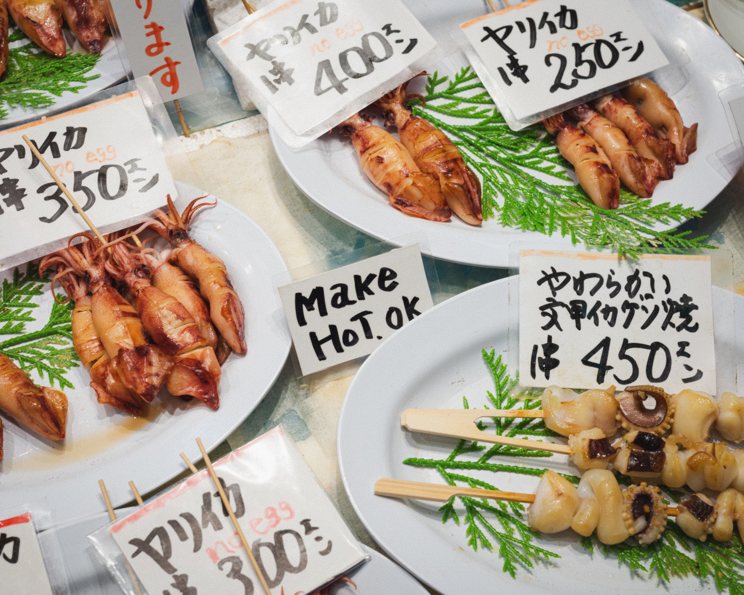 Fresh squid on plates with handwritten Japanese and English signs indicating prices and descriptions. Some signs say "Make Hot. OK" and "450 yen". The squid are garnished with green leaves and presented in a market or street food setting.