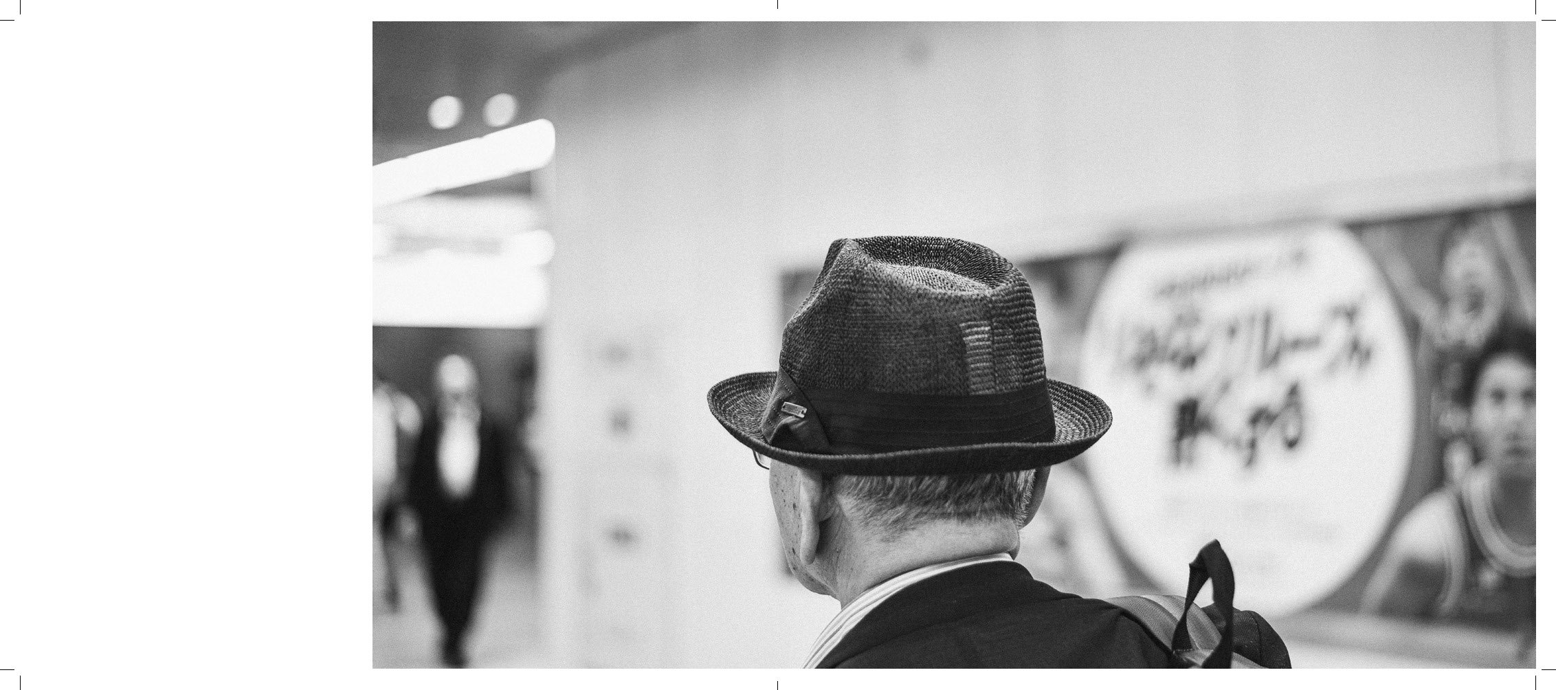 Back view of a person wearing a fedora hat and a suit, standing in an indoor setting with banners and people in the background.