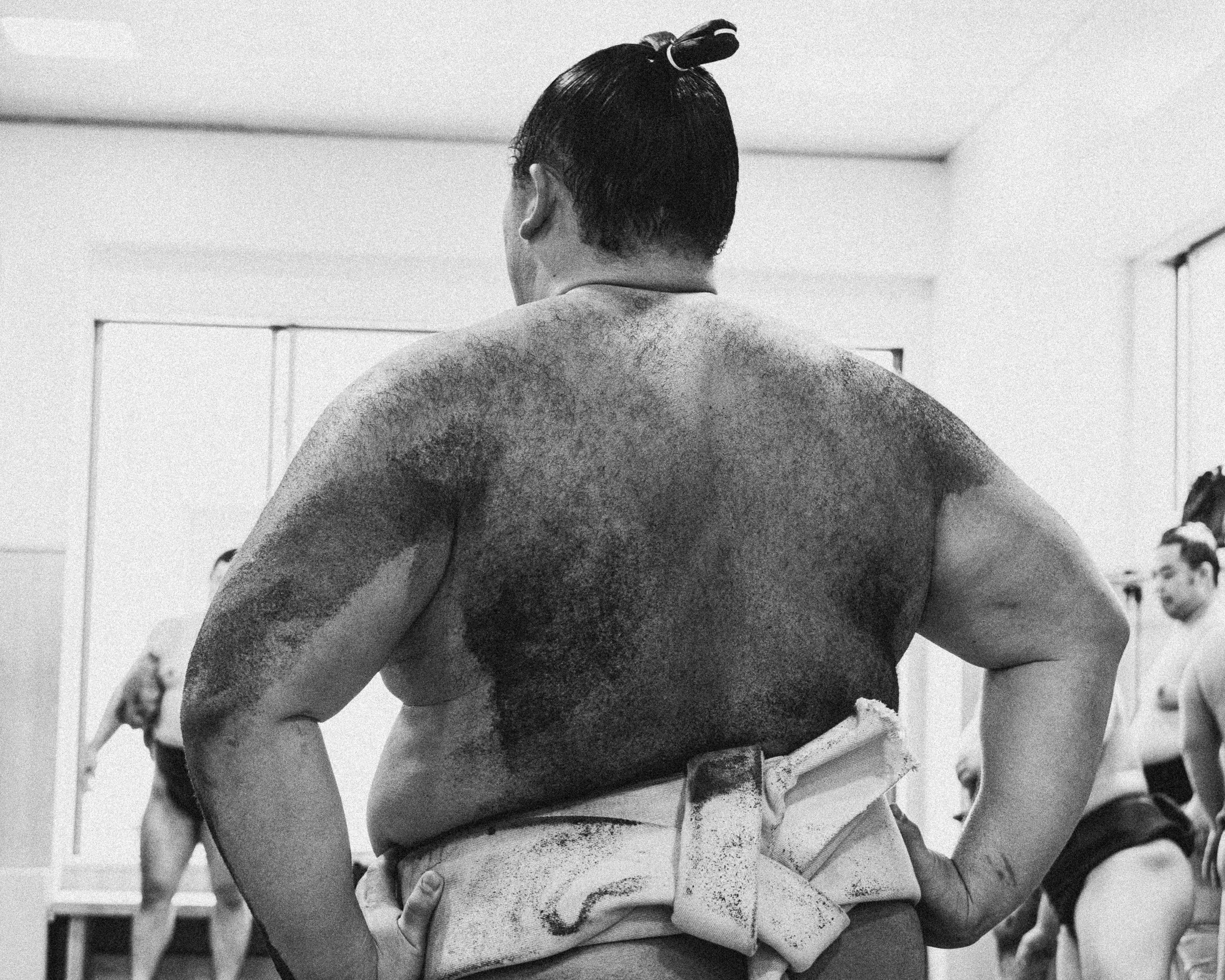 Back of a sumo wrestler with a topknot, practicing in a dojo, with other wrestlers in the background, black and white photo