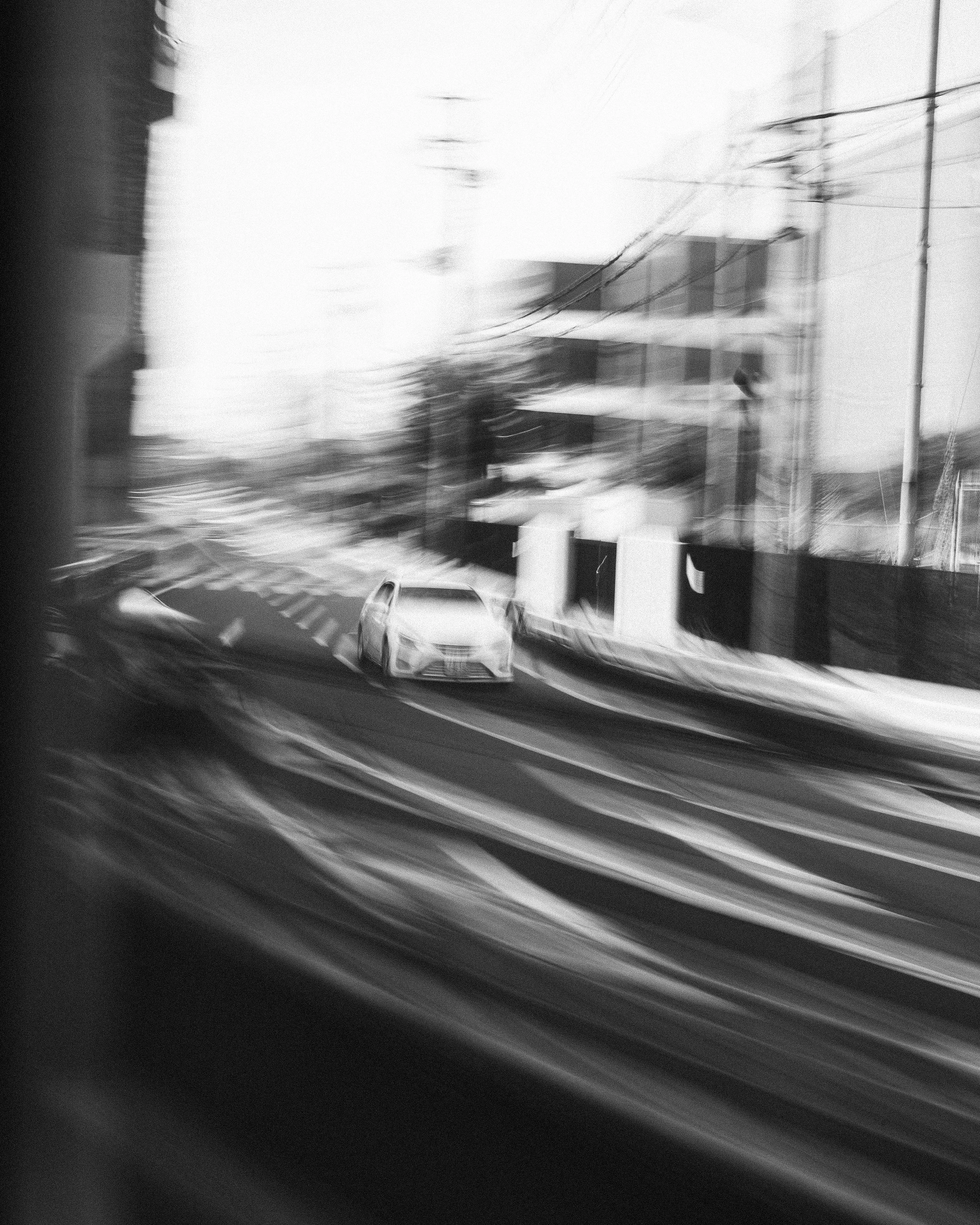 Black and white photo taken from a moving vehicle showing a blurred city street, white car, buildings, power lines, and road markings.