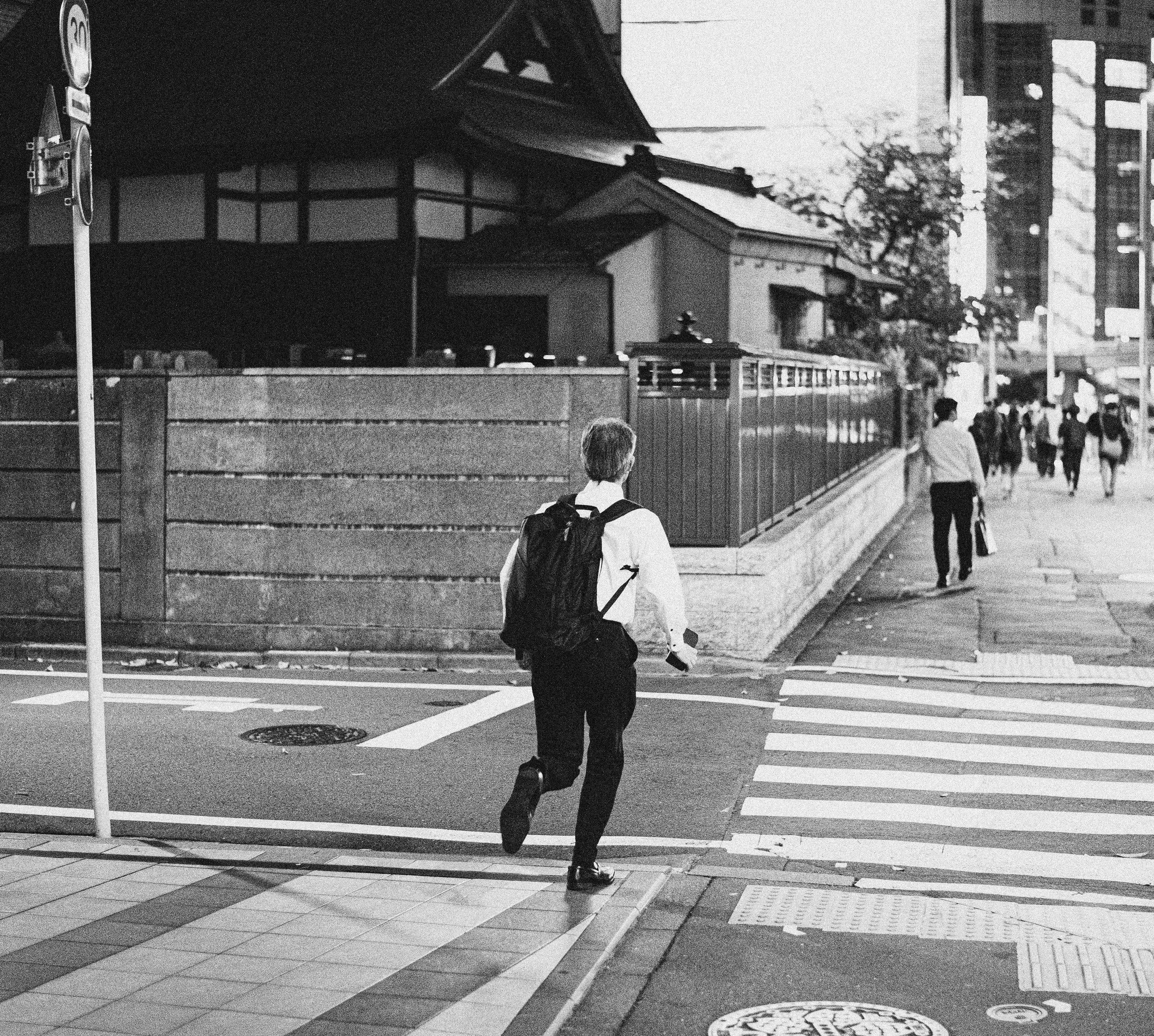 A man with a backpack crossing a city street at a crosswalk, with pedestrians and buildings in the background.