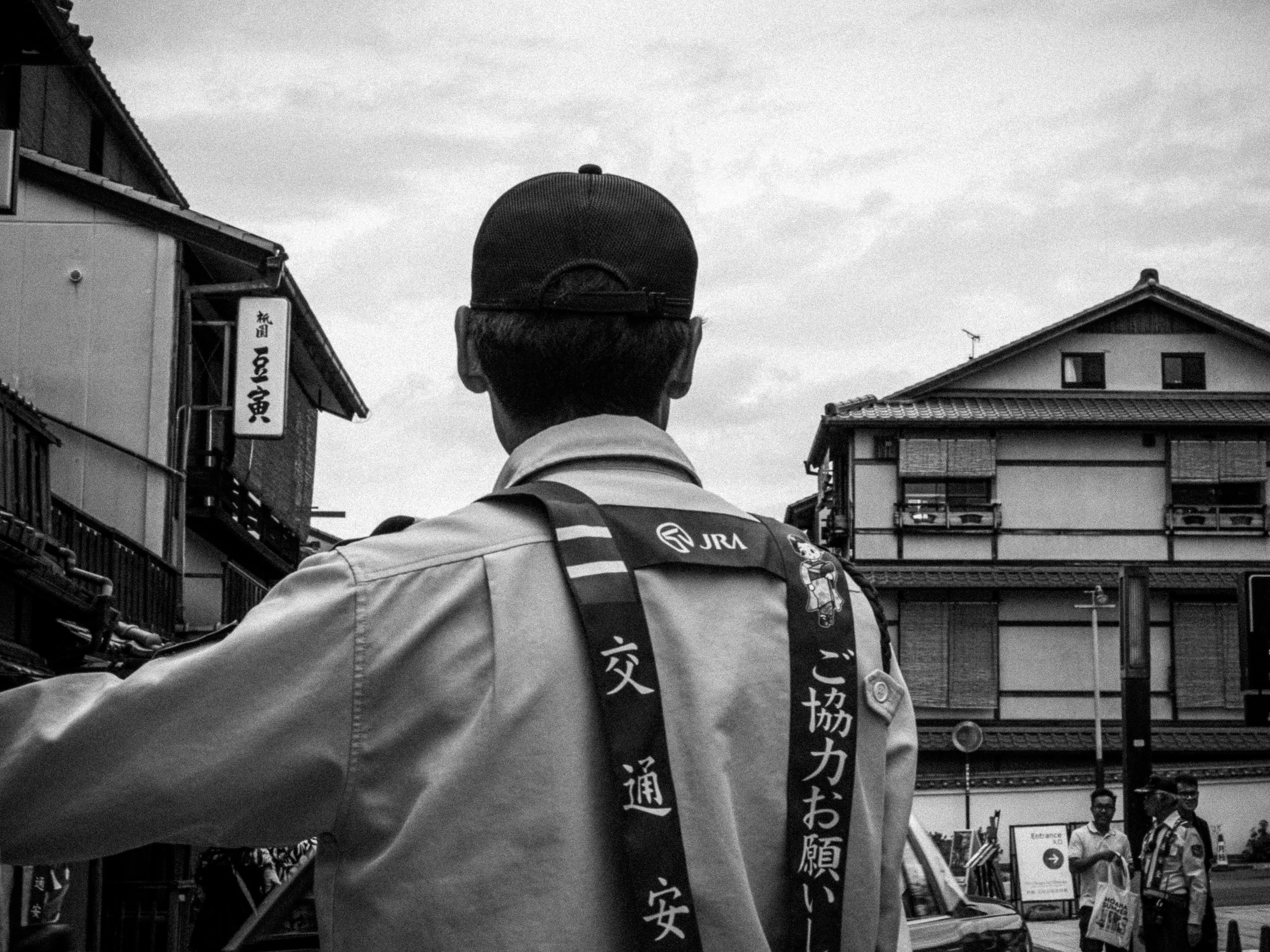 A man wearing a cap and a jacket with Japanese writing and logos faces away, standing on a street with traditional Japanese buildings, some pedestrians, and street signs in the background.