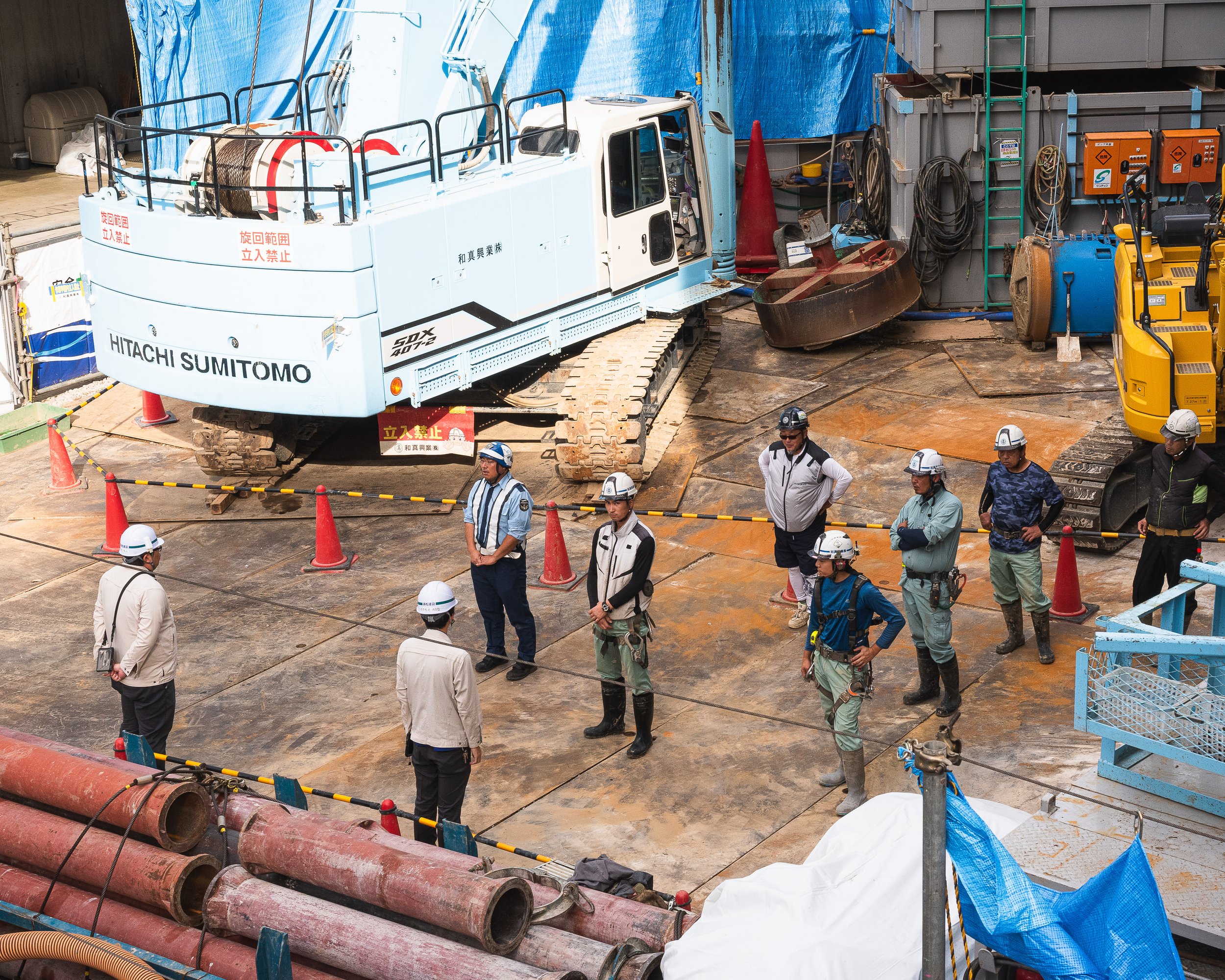 Construction site with workers in helmets and safety gear standing in a circle, a large Crawler crane with the label "HITACHI SUMITOMO" in the background, pipes, and construction equipment visible.