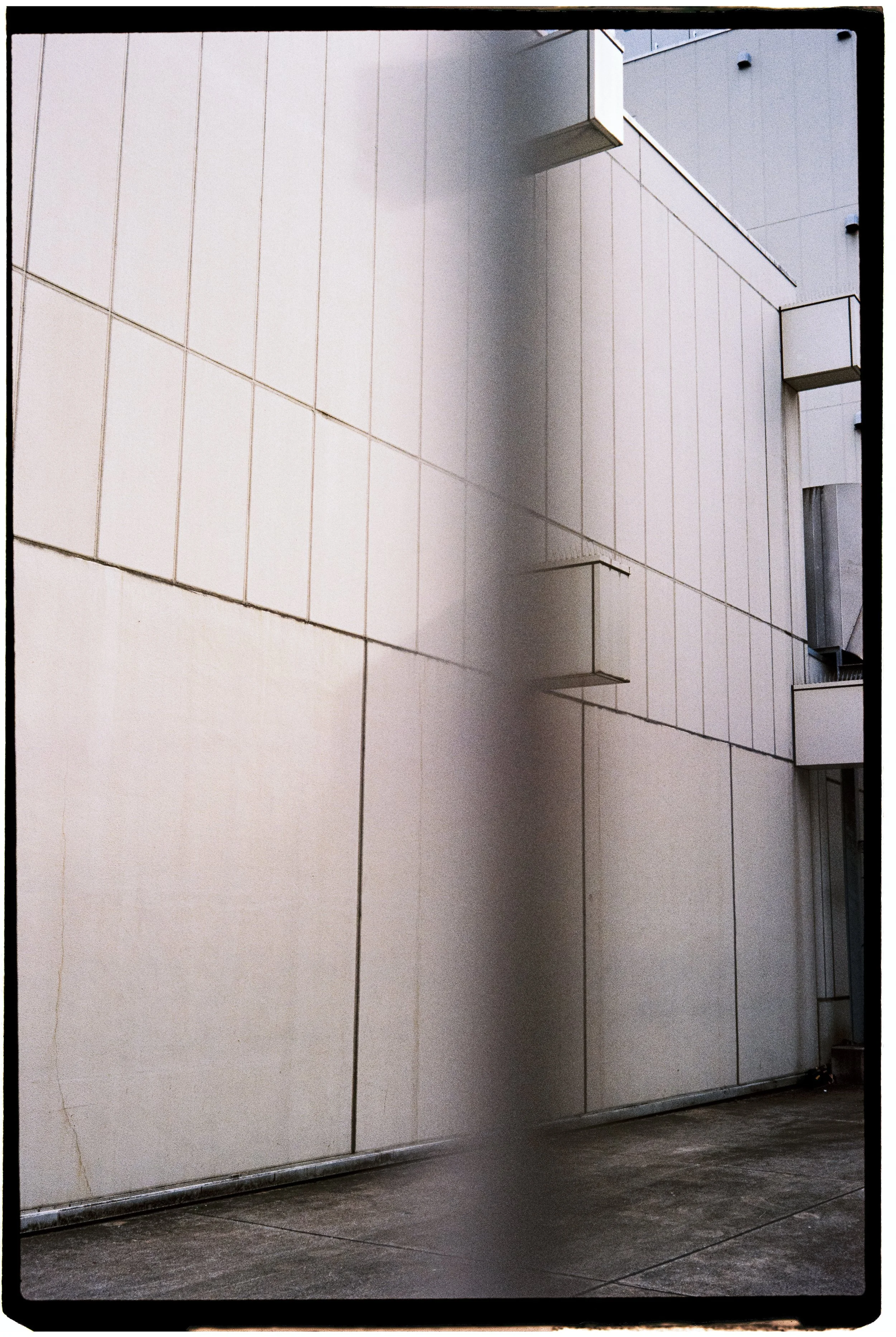 A modern, white tiled wall with small protruding vents and a small overhang. The wall meets a concrete floor, and the photo is taken at a slight upward angle.