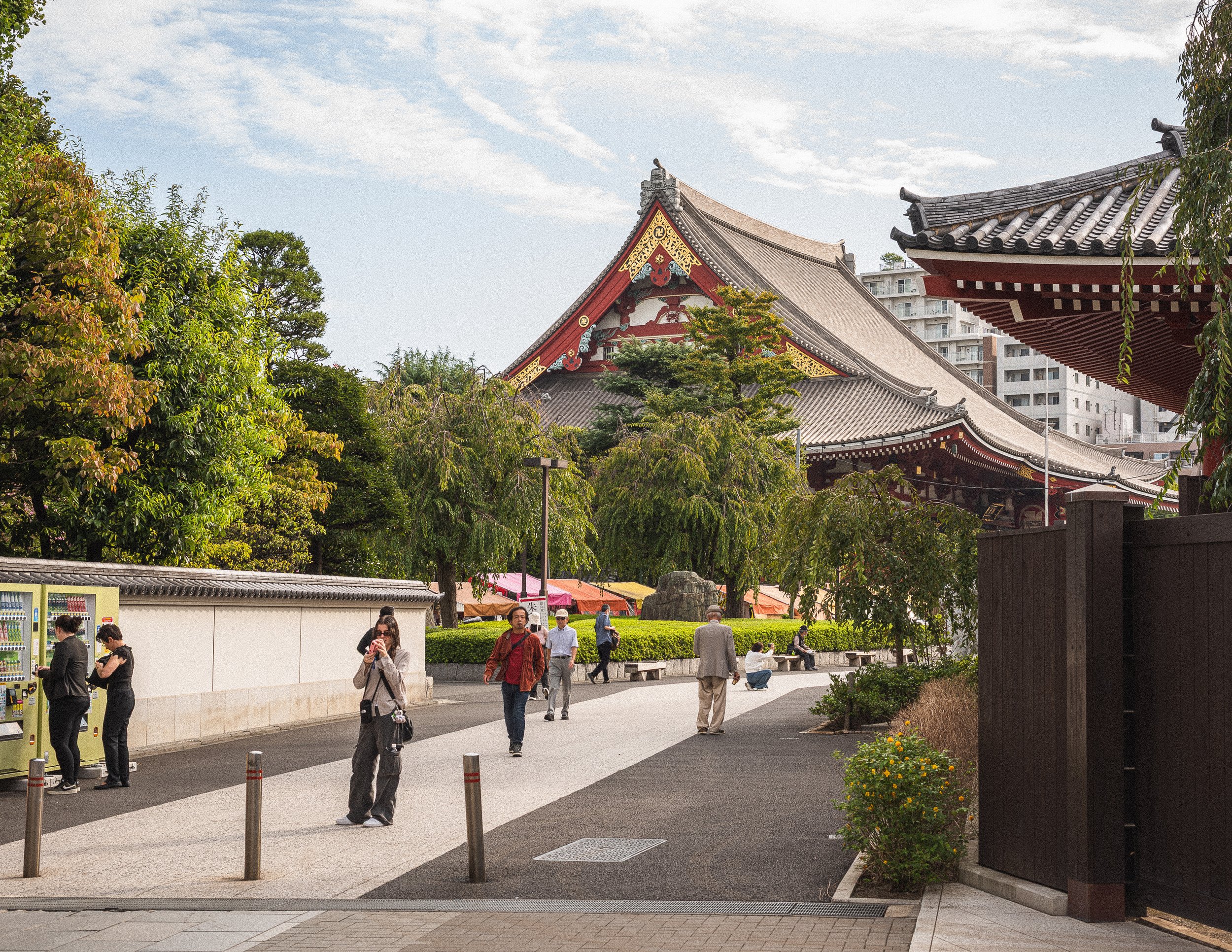People walking outside a traditional Japanese temple with trees and a modern building in the background.