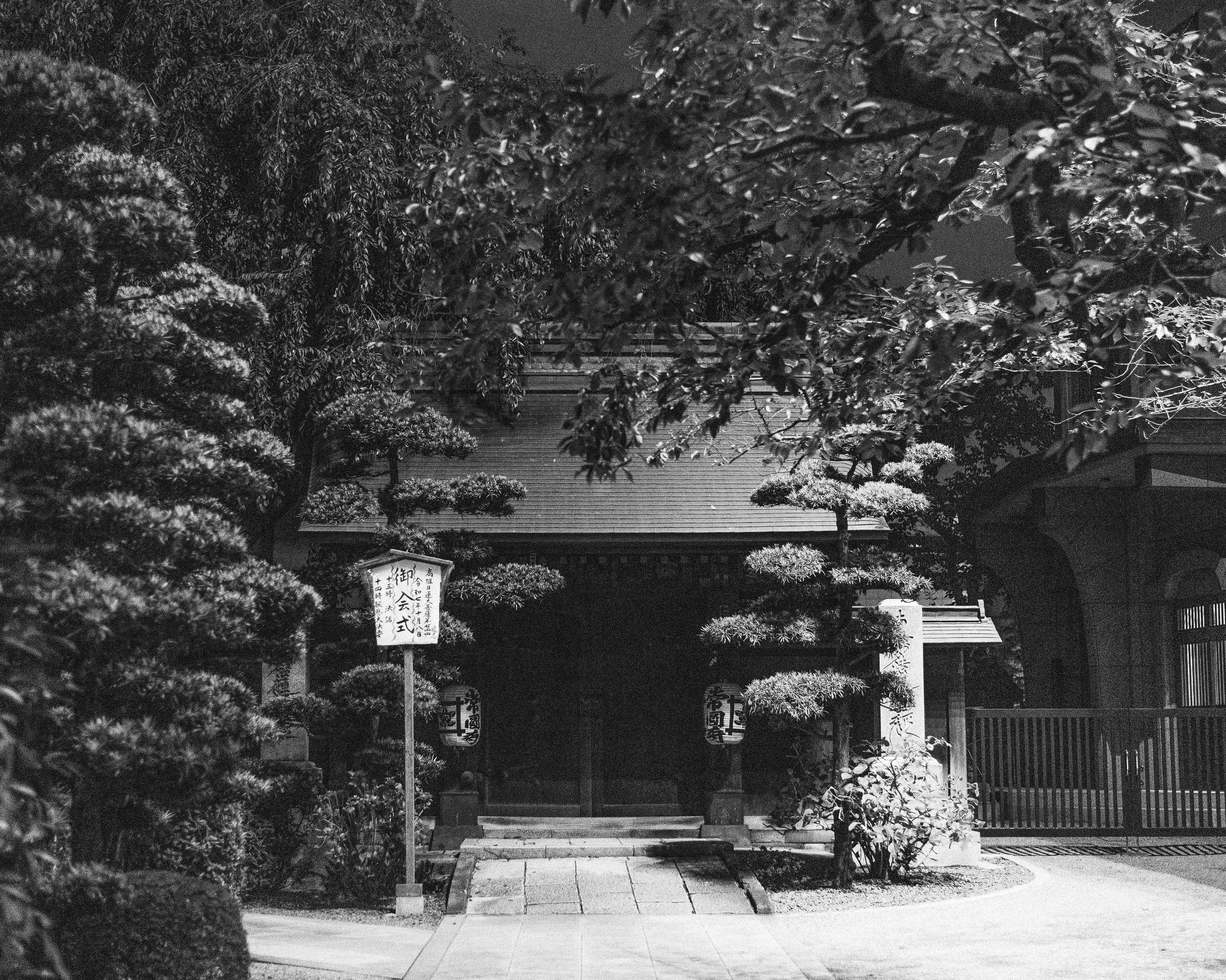 A black and white photo of a small traditional Japanese shrine or temple entrance, surrounded by manicured bushes, trees, and a pathway leading to the entrance. There are paper lanterns and a sign with Japanese characters near the entrance.