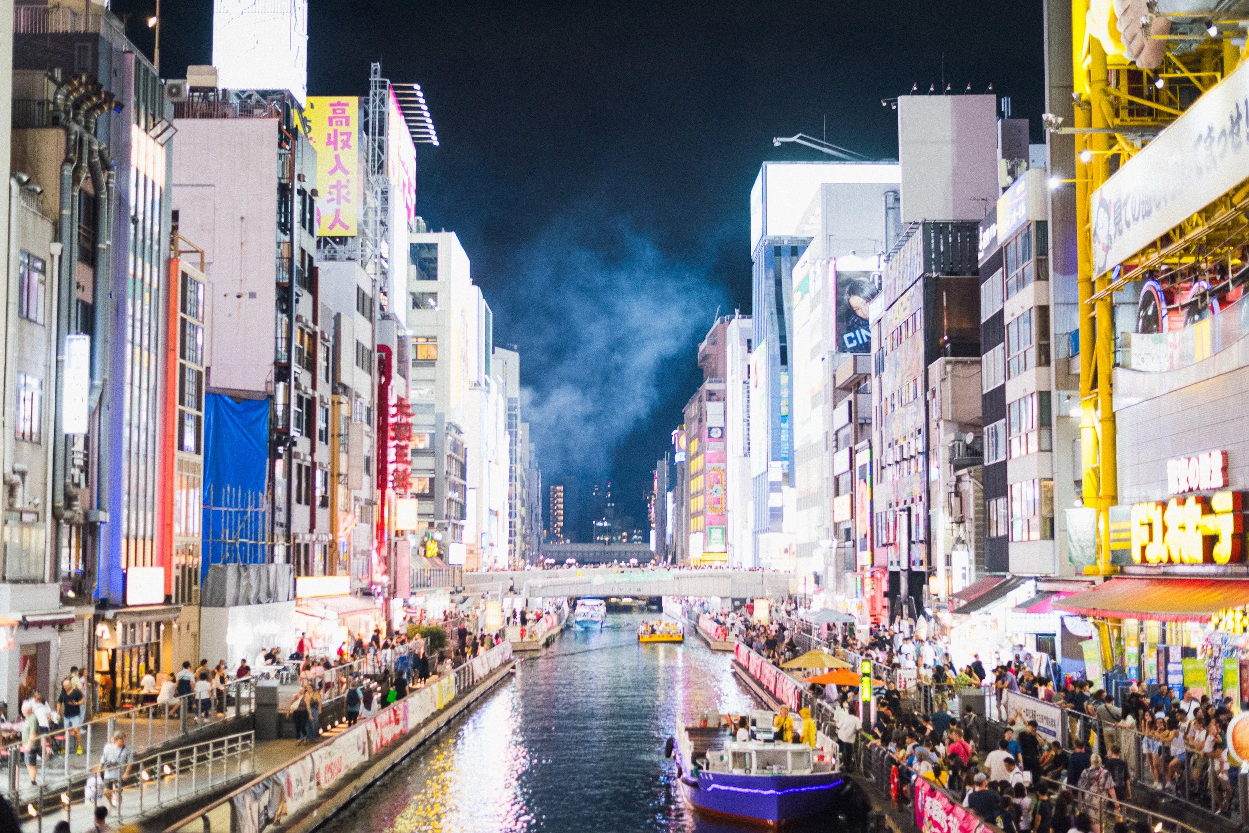 Night scene of a canal in an urban area with tall, brightly lit buildings on both sides, crowded with people, some walking on the sidewalks and others on boats on the water.