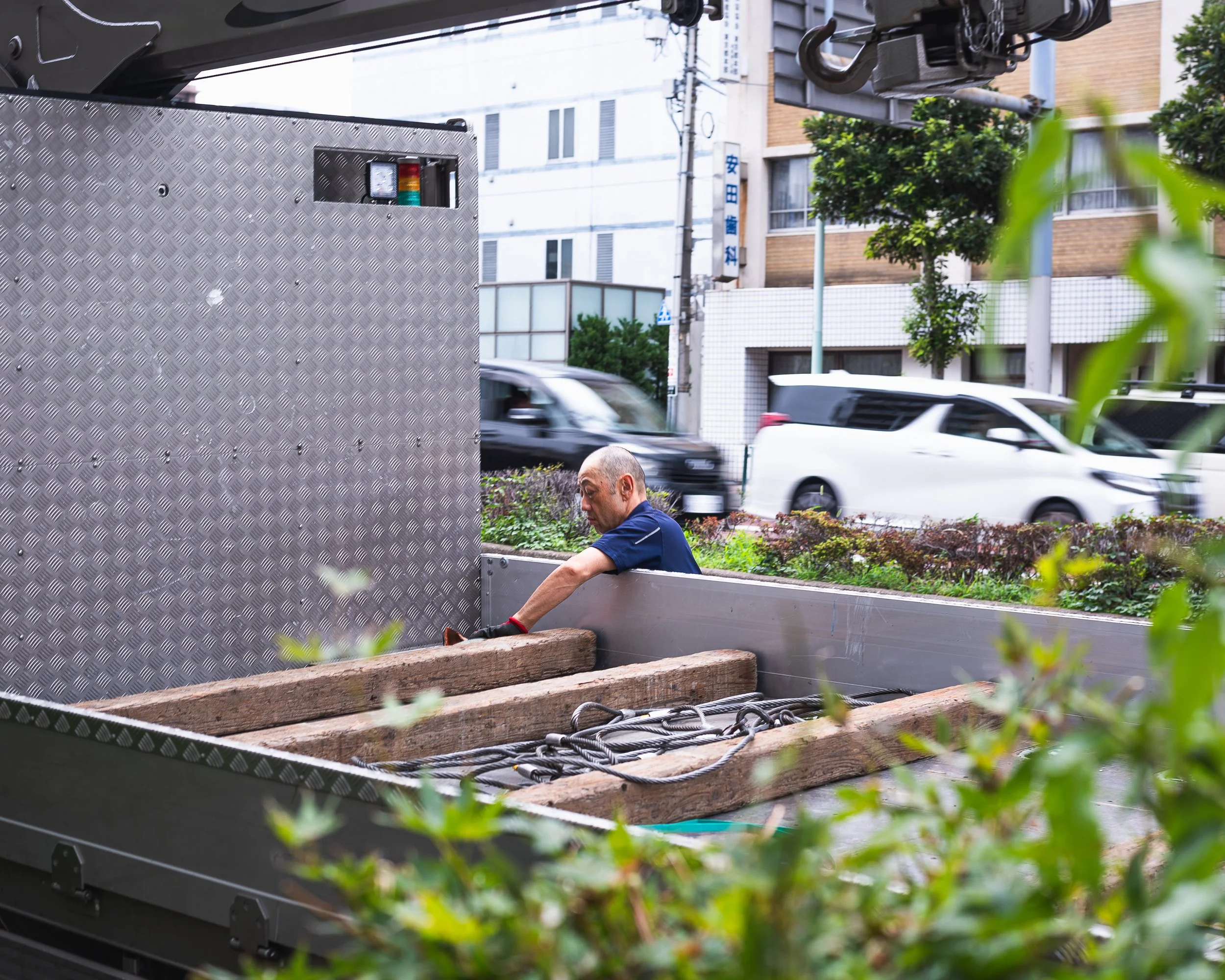 A man sitting on a truck bed among large wooden planks and ropes in an urban street setting with buildings, cars, and trees in the background.