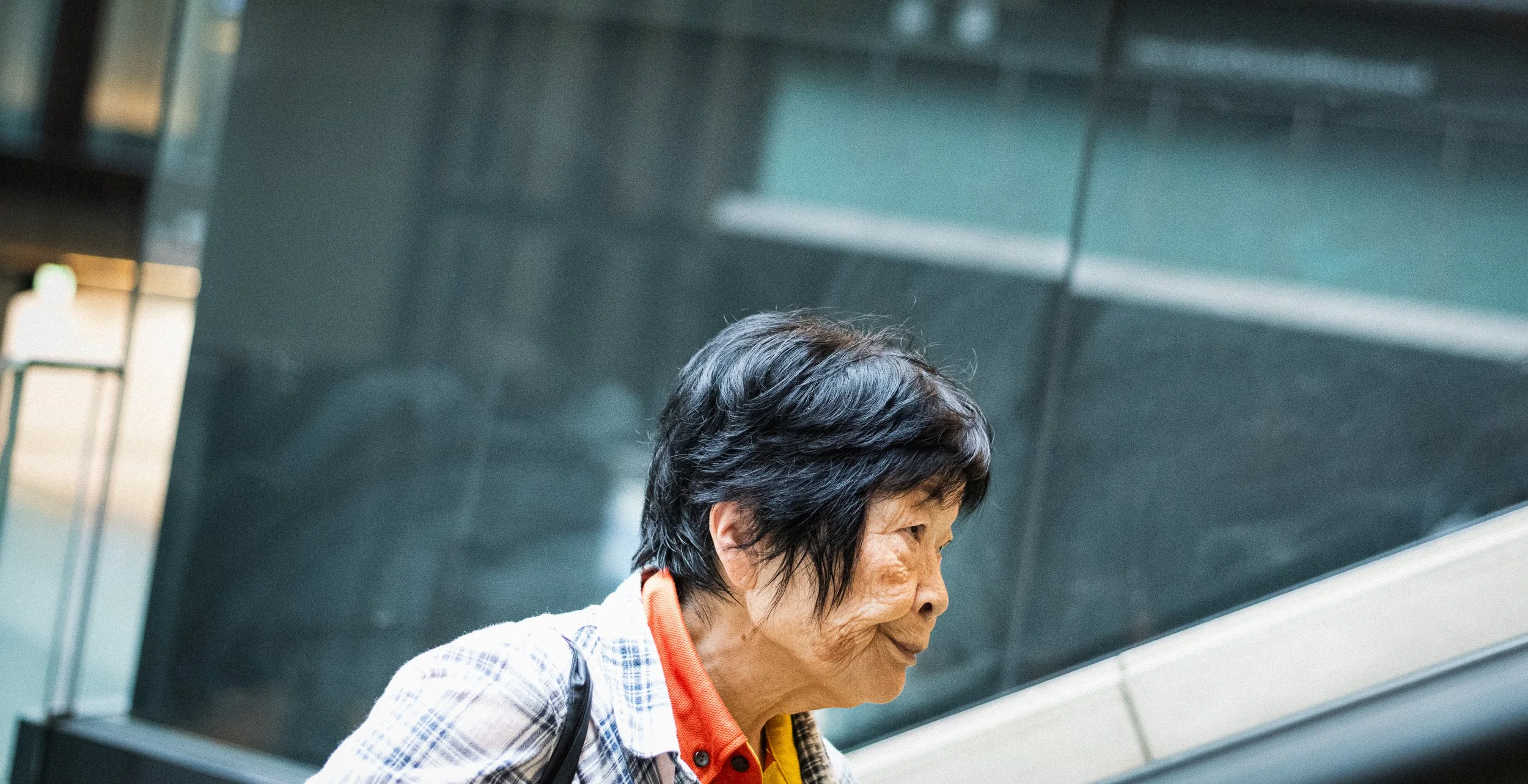 Side view of an elderly Asian woman with short black hair, wearing a checkered jacket and a red shirt, standing indoors near a shiny, dark gray wall.
