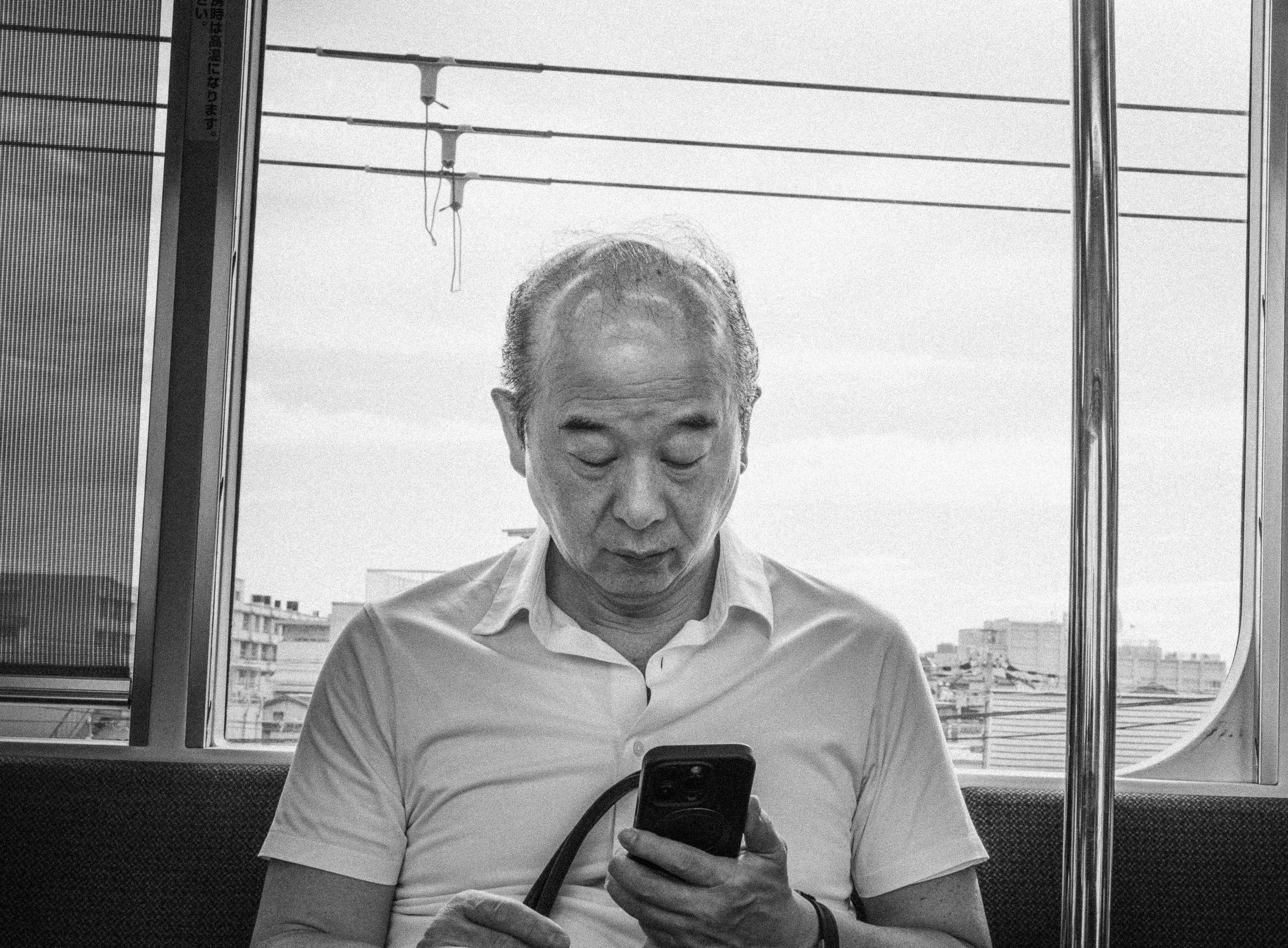 An older man with a bald head and white shirt sitting inside a train, looking down at his smartphone.