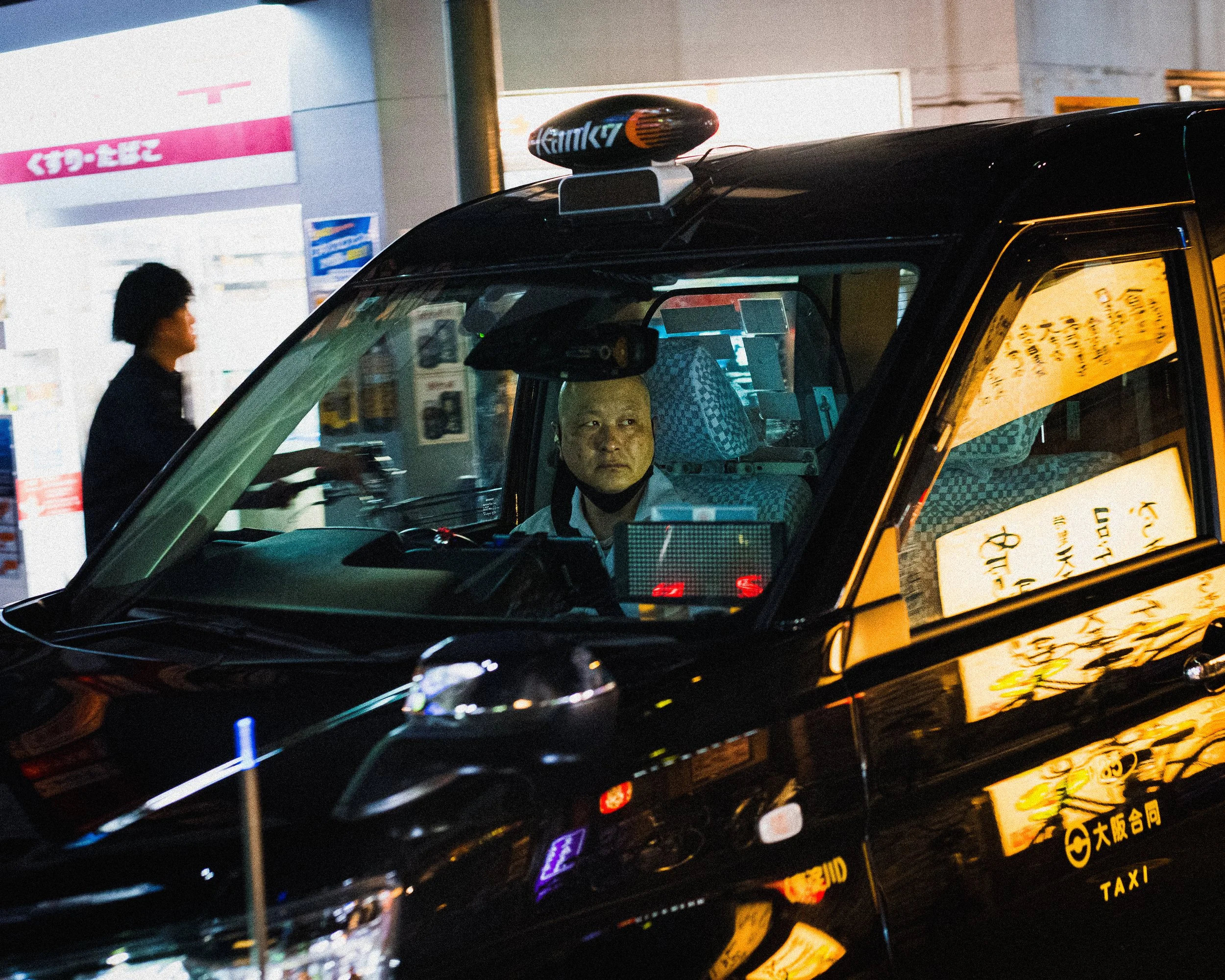 A man with a shaved head and a face mask sitting inside a black taxi at night, with illuminated signs reflected on the car's surface in an urban setting.