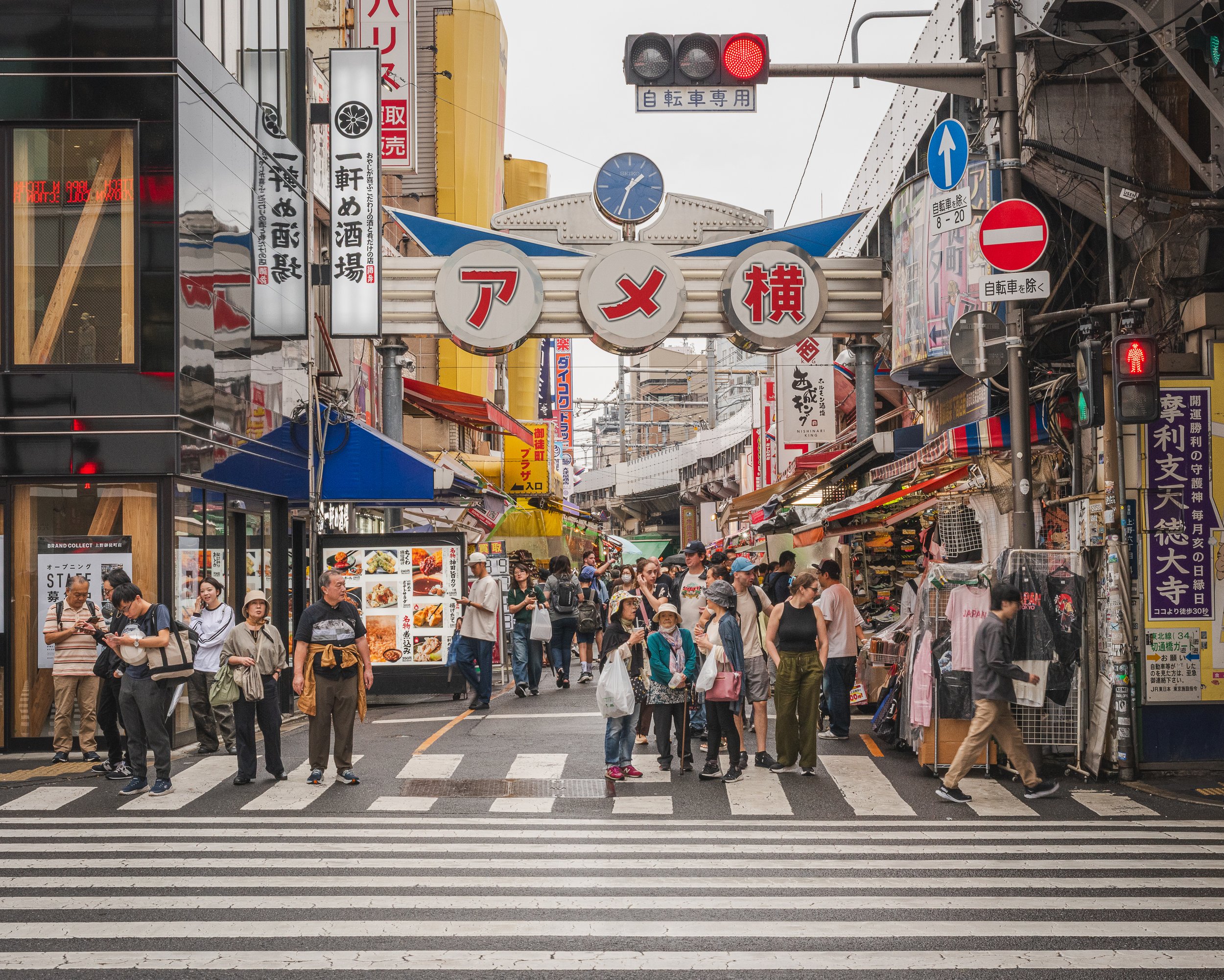 Crowd of people walking and shopping on a busy street in Japan with signs and storefronts in Japanese, including a large archway with red and white Japanese characters and a clock above.