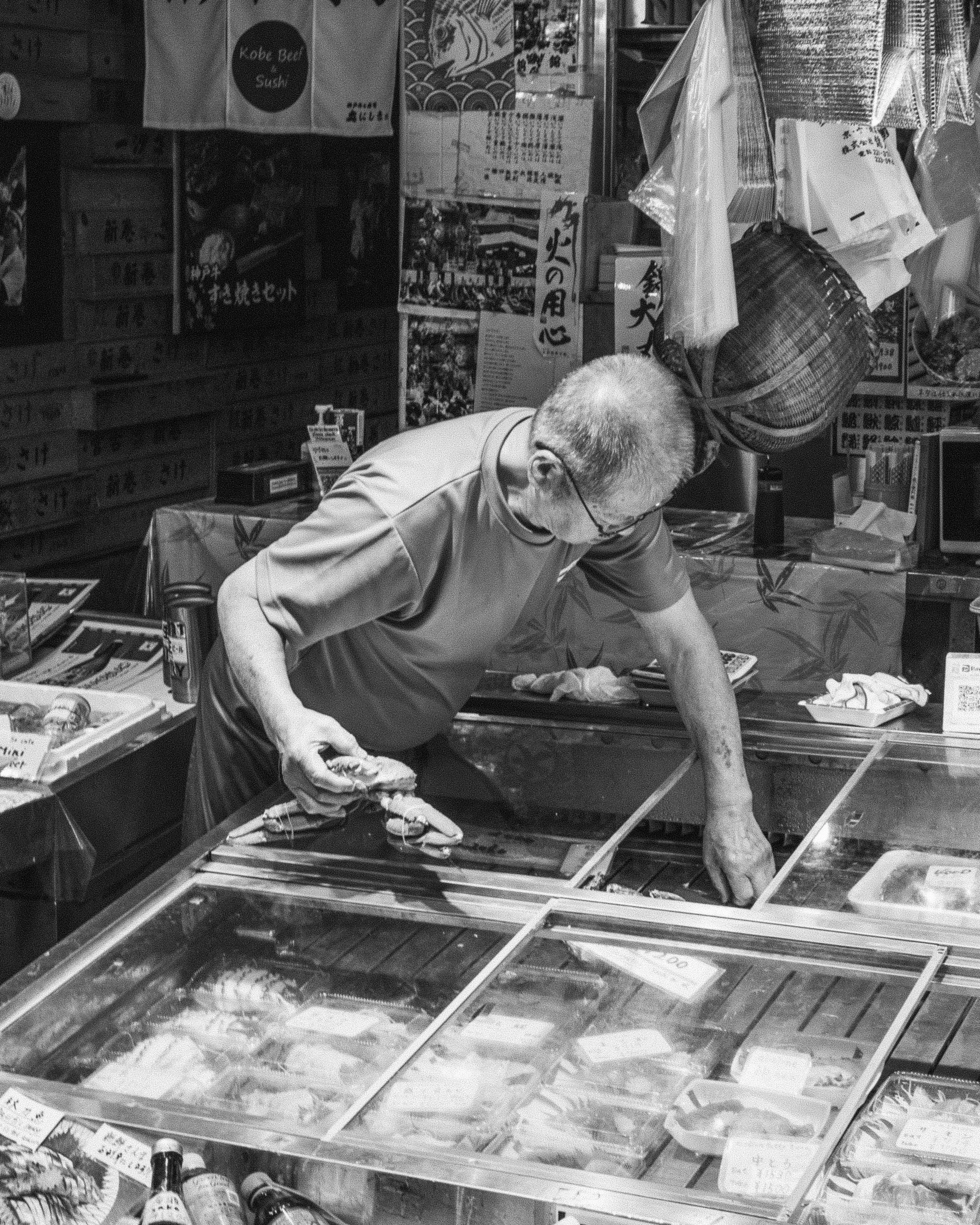 An elderly man with glasses is working at a seafood stall, holding a lobster and arranging seafood in a glass display case.