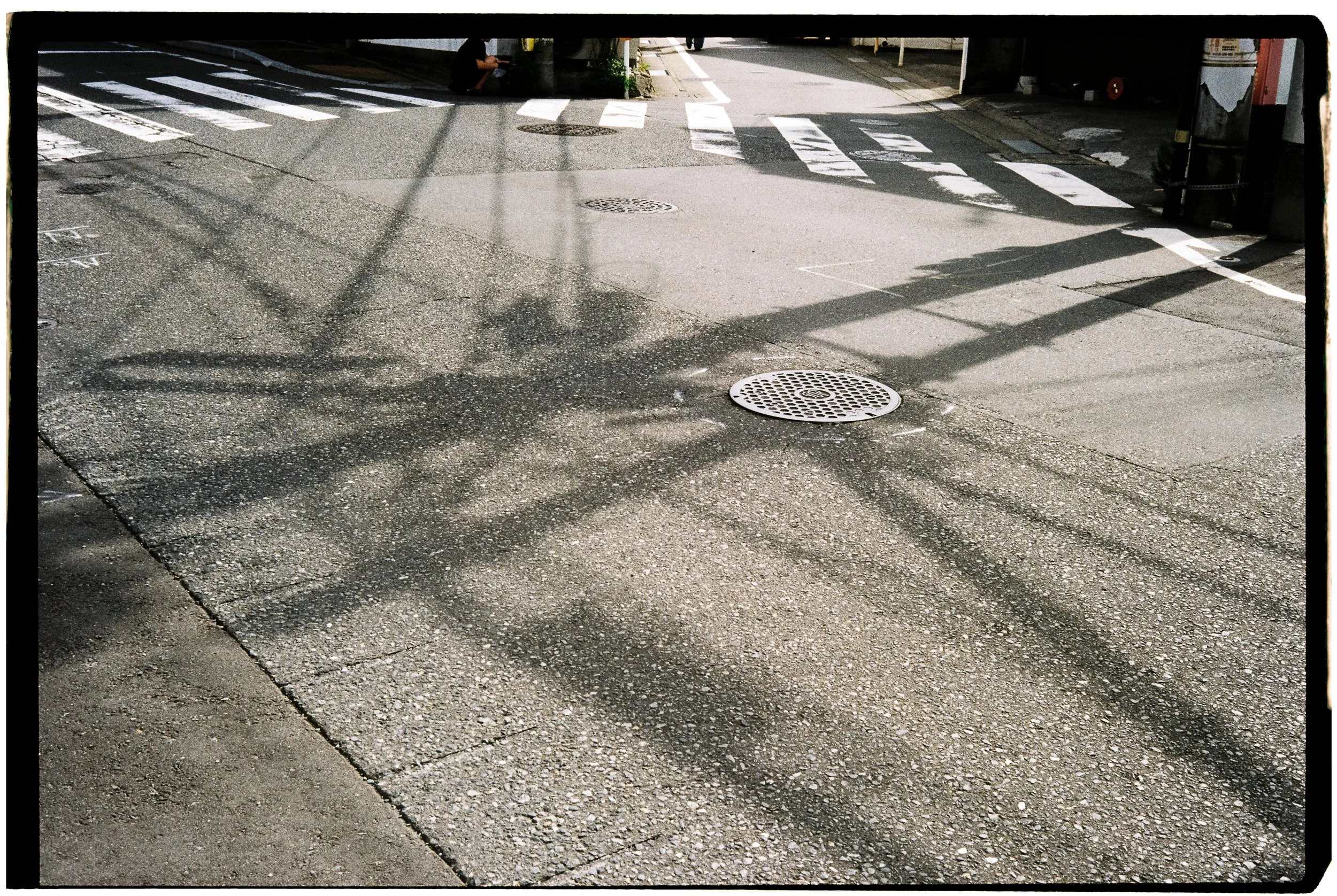 Shadows of power lines cast across an urban street with a manhole cover, crosswalk lines, and a person sitting at the curb.