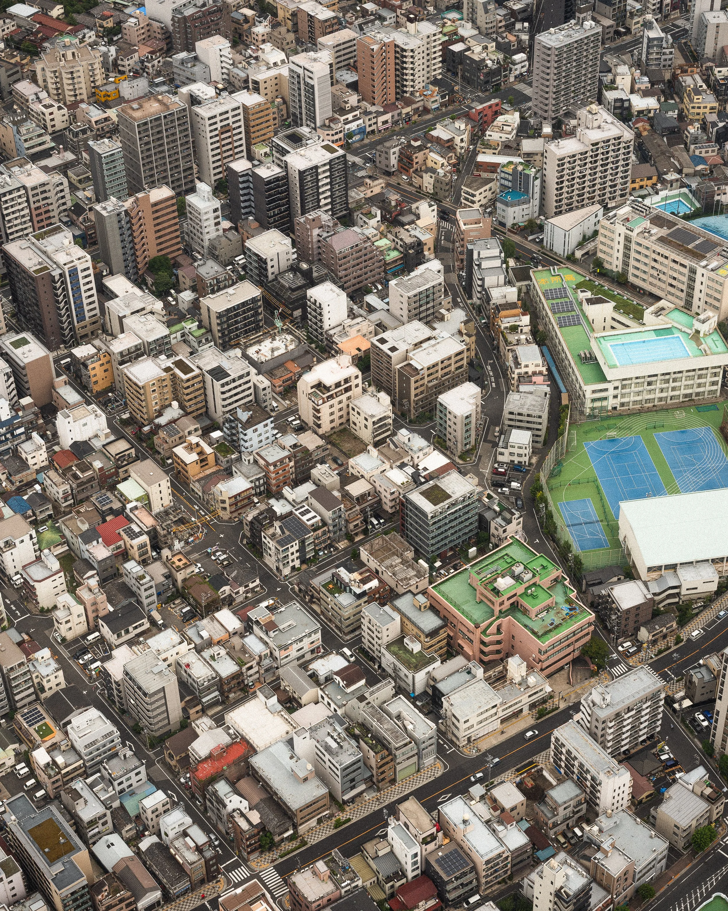 Aerial view of a densely populated urban city with numerous high-rise and low-rise buildings, streets, and sports courts surrounded by green fencing.